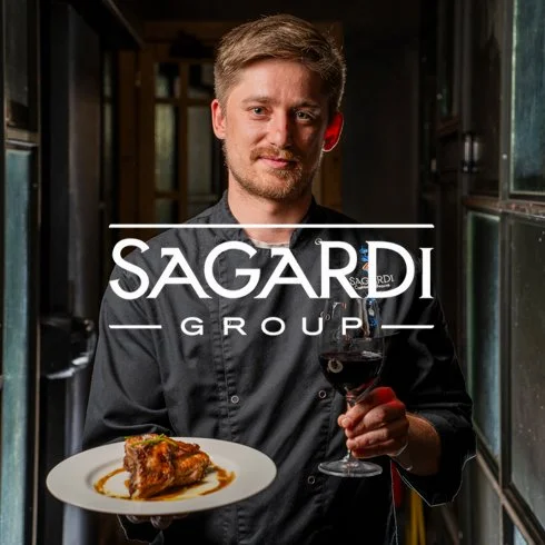 Homem segurando uma taça de vinho tinto e um prato de comida em um ambiente de restaurante.