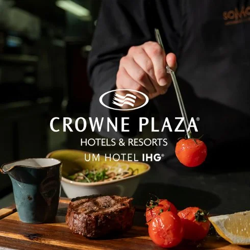 Pessoa segurando um espetinho de tomate cereja com um garfo em uma tábua de madeira, com uma peça de carne, tomates e uma fatia de limão ao lado, em um ambiente de restaurante.