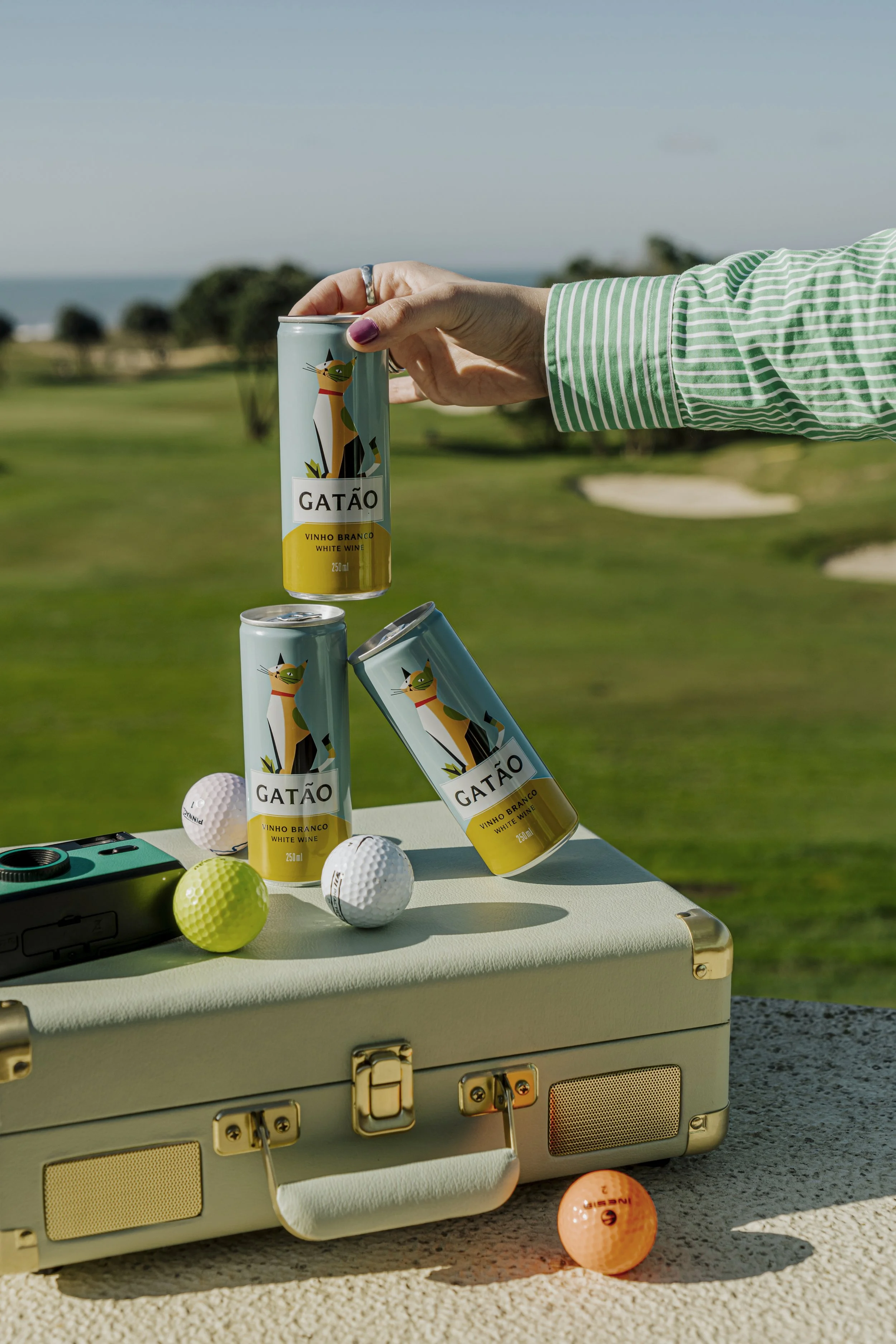 Mãos segurando uma lata de refrigerante ao lado de outras latas, em um cenário ao ar livre com grama e céu azul, ao lado de bolas de golfe e um equipamento de golfe.