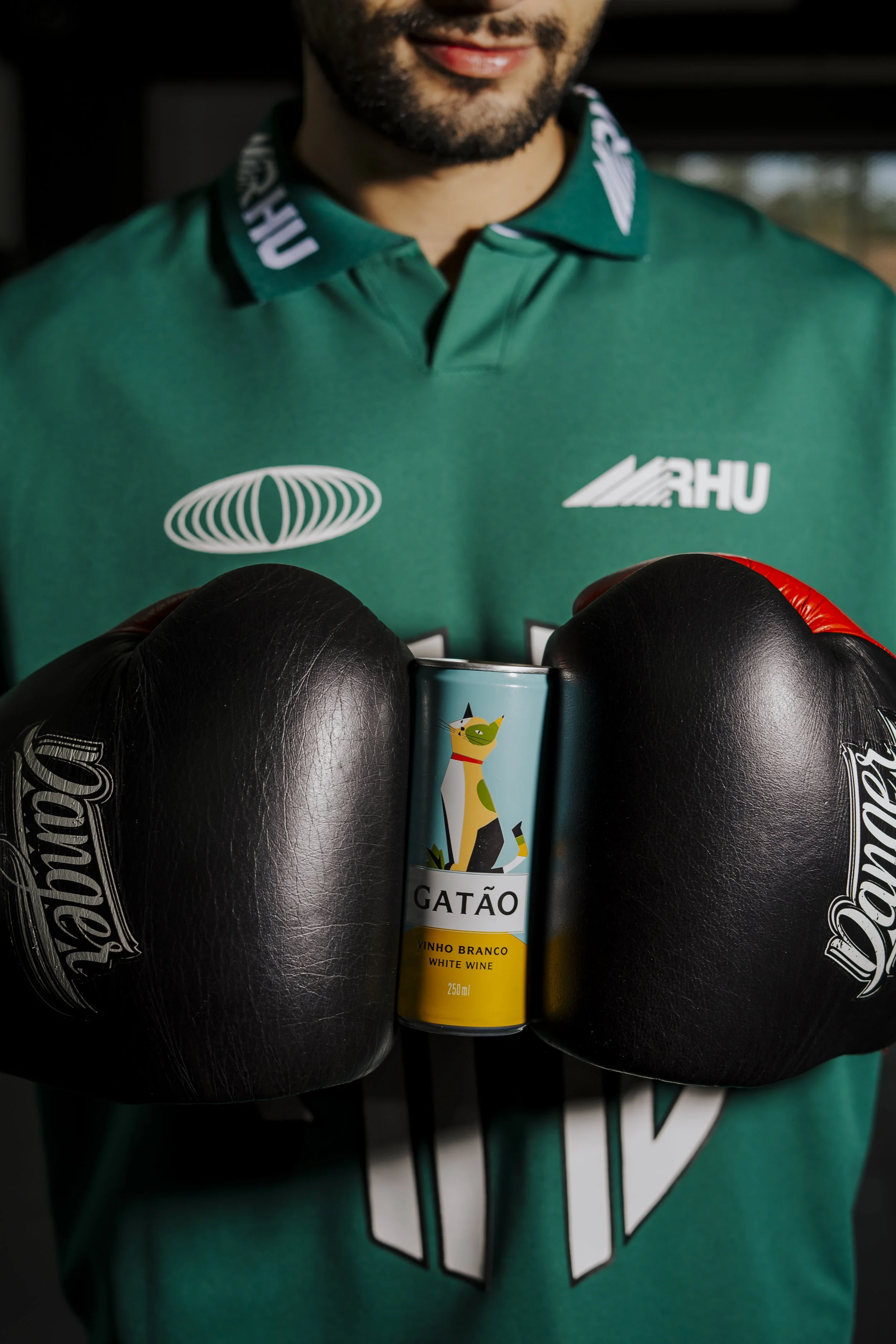 Homem com barba usando camiseta verde com logos, segurando uma latinha de vinho branco decorada com uma ilustração de gato e duas luvas de boxe negras.