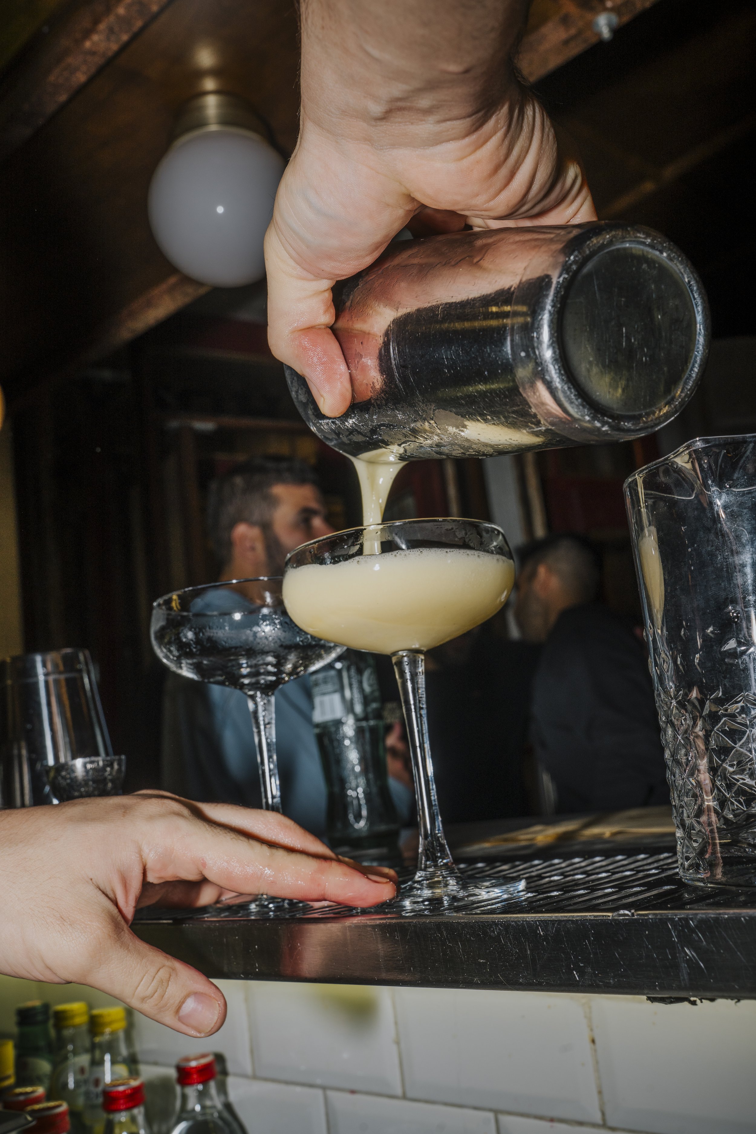 Pessoa despejando uma bebida de um copo em uma taça de cocktail, enquanto outra pessoa segura a taça. A cena ocorre em um bar com pessoas ao fundo.