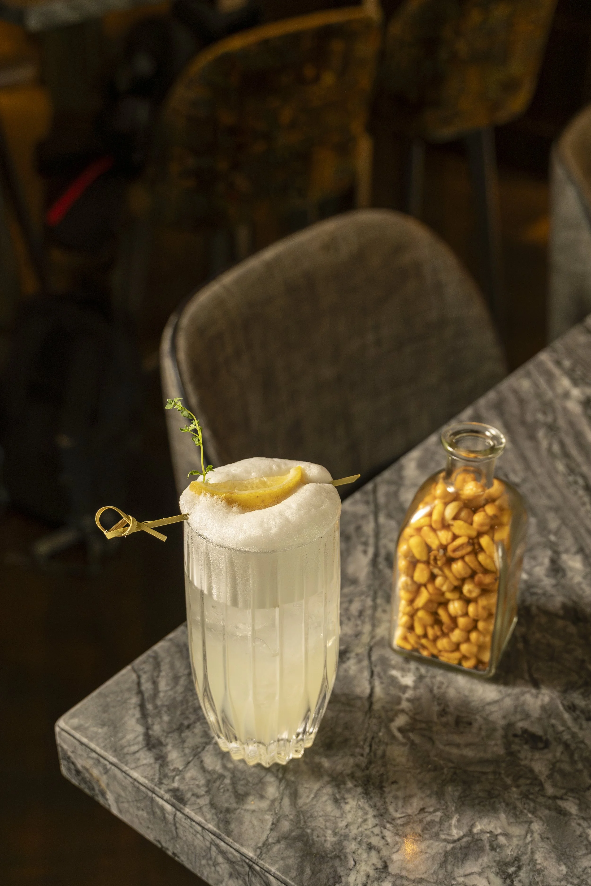 Copo de coquetel com espuma, decorado com uma fatia de limão e um raminho de erva, ao lado de um pote com amendoins, sobre uma mesa de mármore.