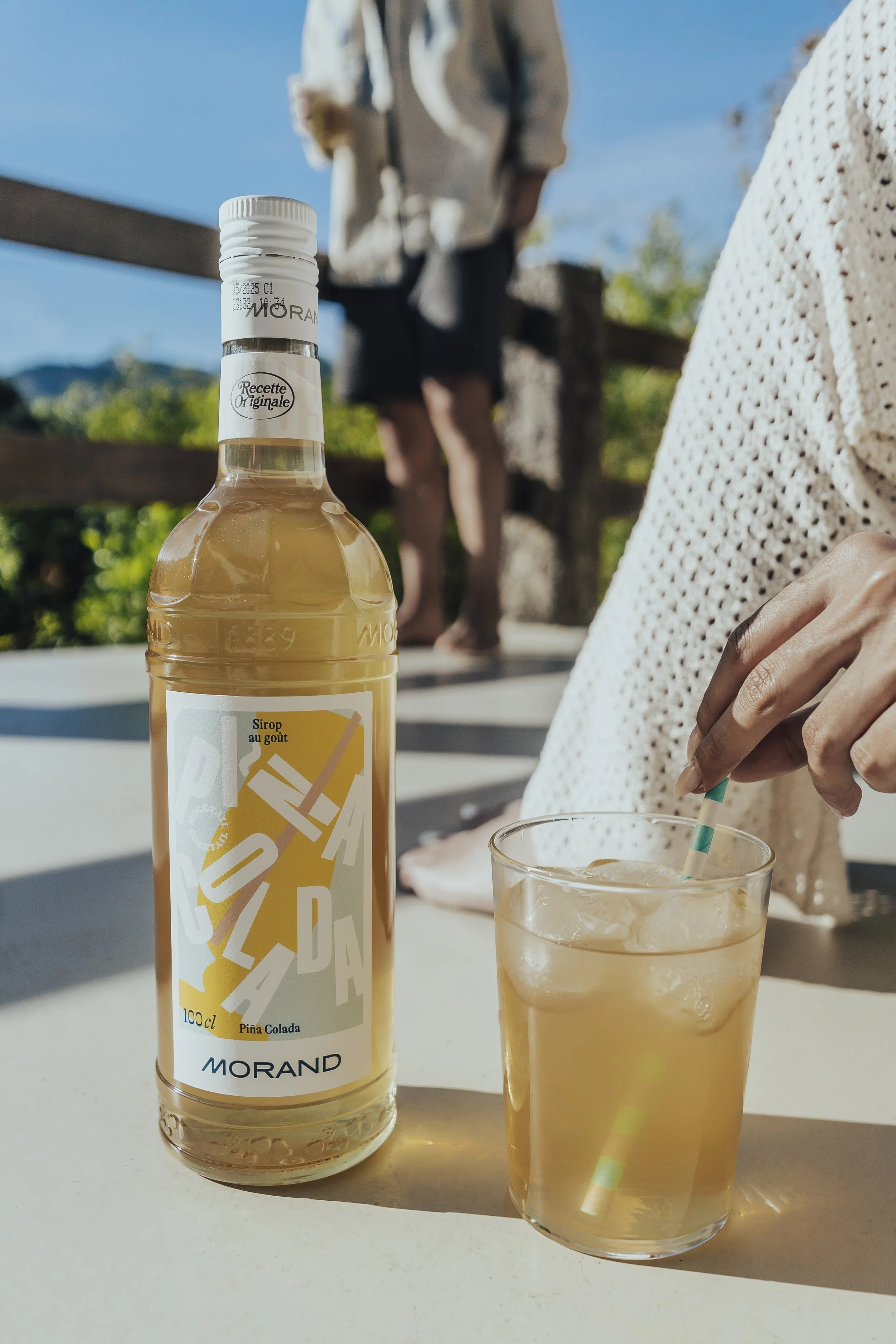 Garrafas de refrigerante de sabor abacaxi e copo de vidro com bebida, gelo e canudo, ao ar livre com pessoas ao fundo.