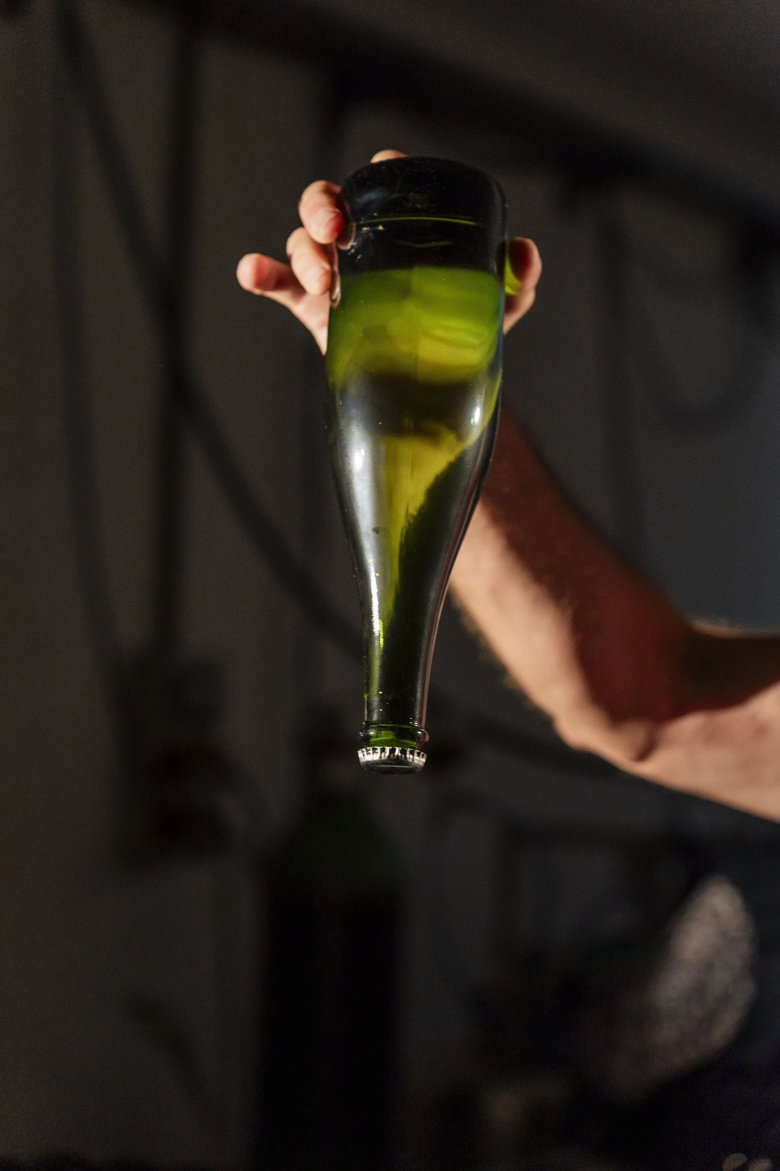 Pessoa segurando uma garrafa de vidro preta e verde, inclinada, com líquido verde dentro, em ambiente interno com fundo escuro.