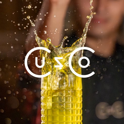 Copo de vidro amarelo com líquido sendo derramado, criando splash na frente de uma pessoa desfocada ao fundo.