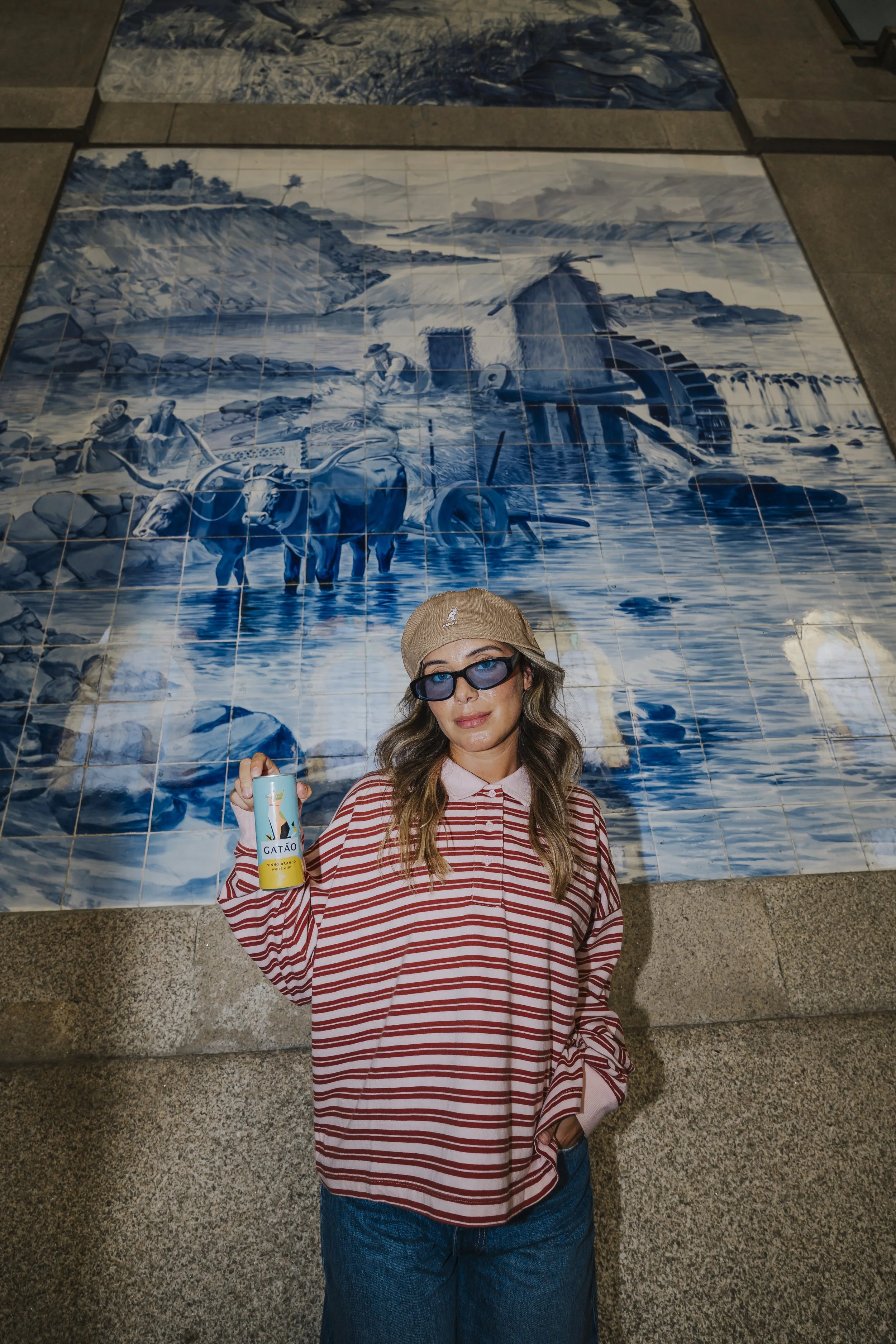 Mulher com óculos de sol, chapéu bege, camiseta listrada vermelha e branca, e jeans, segurando uma lata de GATÃO, em frente a um mural de azulejos azul e branco que retrata uma cena de pescadores e barcos na água.