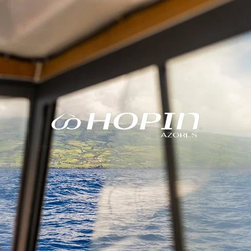 Vista de uma paisagem marítima vista de dentro de uma embarcação, com janelas à frente, mostrando água e terra ao longe, com o texto 'HOPIN AZORES' sobreposto