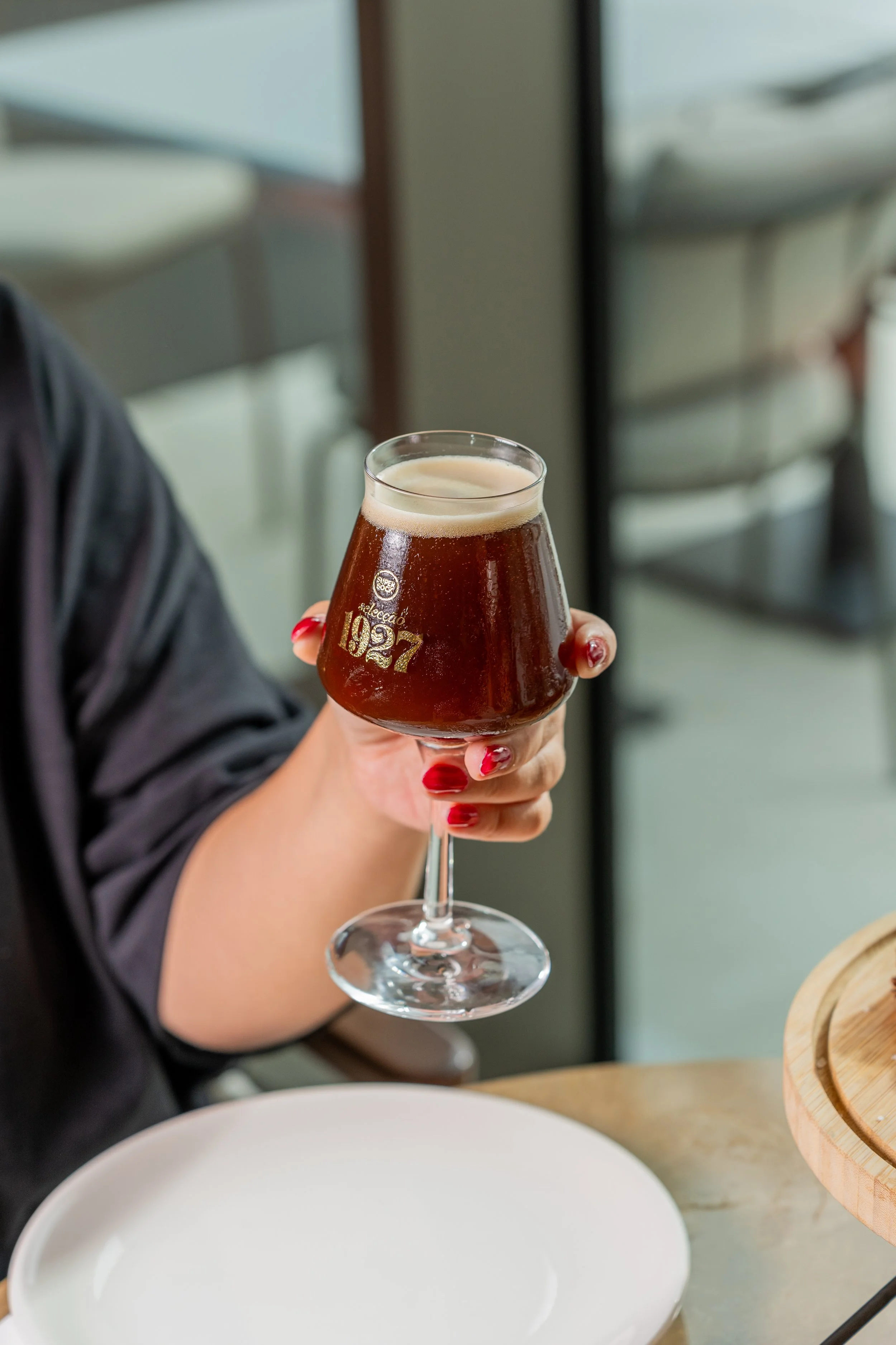 Pessoa segurando um copo de vidro com uma bebida escura e espuma na parte superior, com unhas pintadas de vermelho.