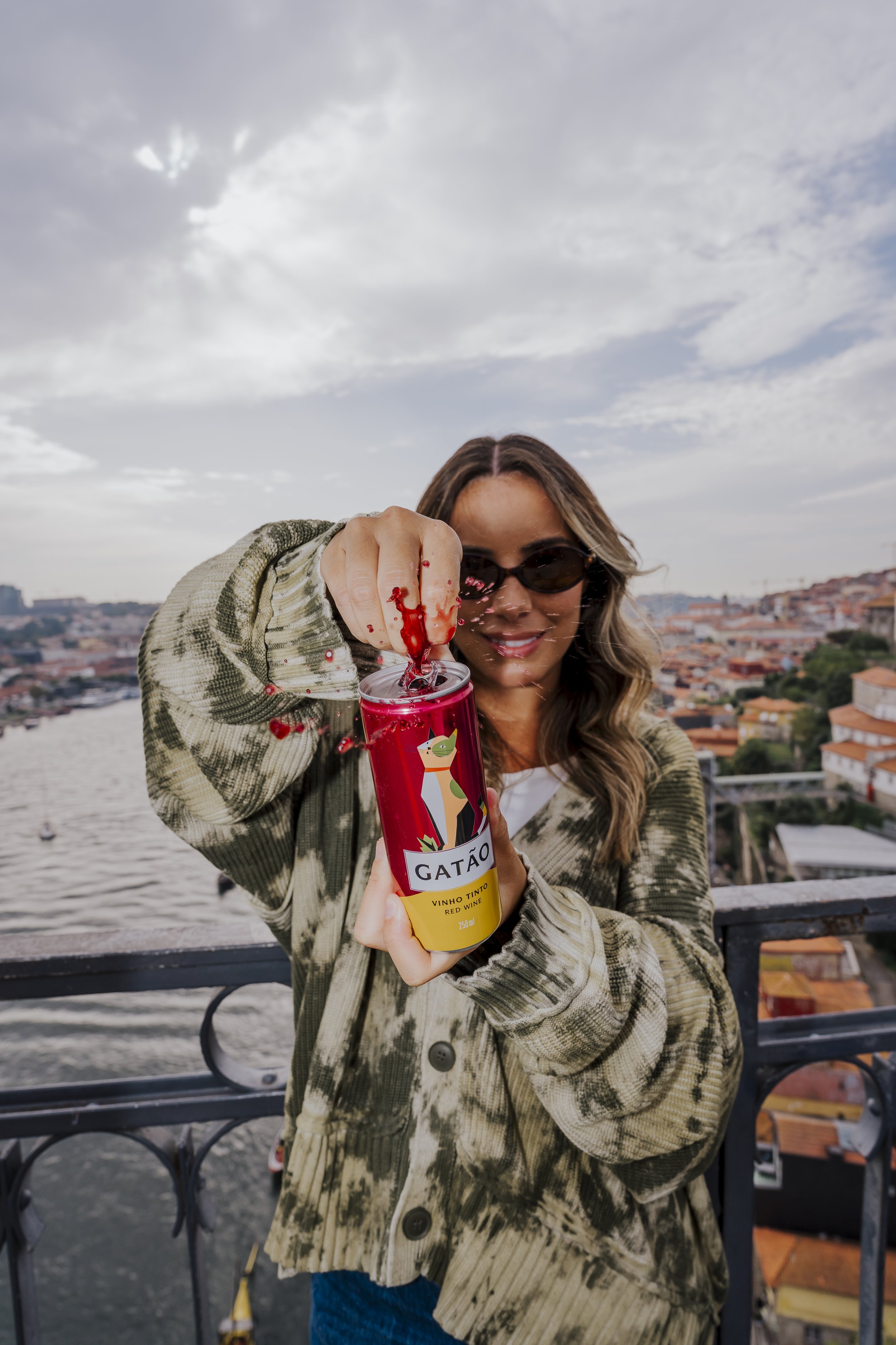 Mulher usando óculos escuros, de cabelo ondulado, com uma jaqueta de camuflagem, segurando uma lata de vinho tinto com uma ilustração de gato, derramando líquido vermelho com uma vista de rio e cidade ao fundo.
