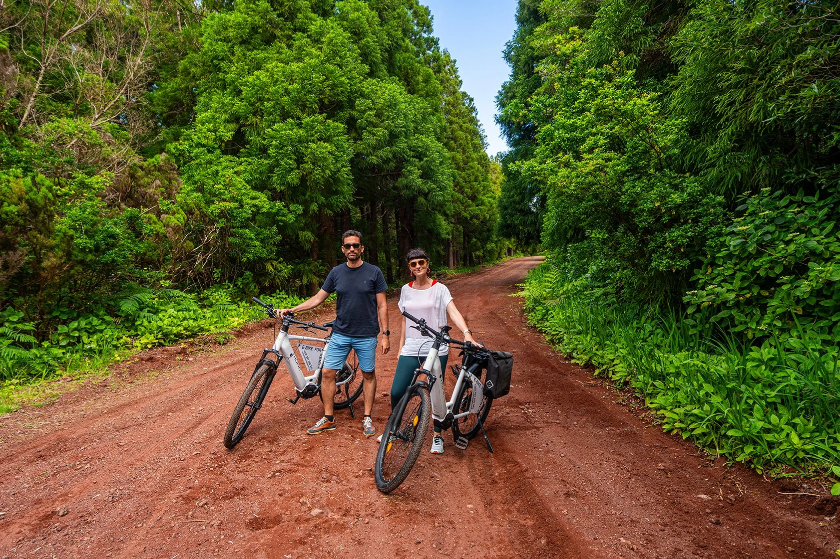 Homens e mulheres com bicicletas elétricas na estrada de terra, rodeados de vegetação verde e árvores ao redor.