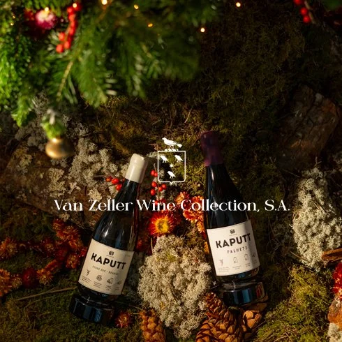 Duas garrafas de vinho Kaputt, uma ao lado da outra, sobre uma superfície coberta de musgo, com bolas de Natal, pinhas e árvores de Natal decorando ao fundo.