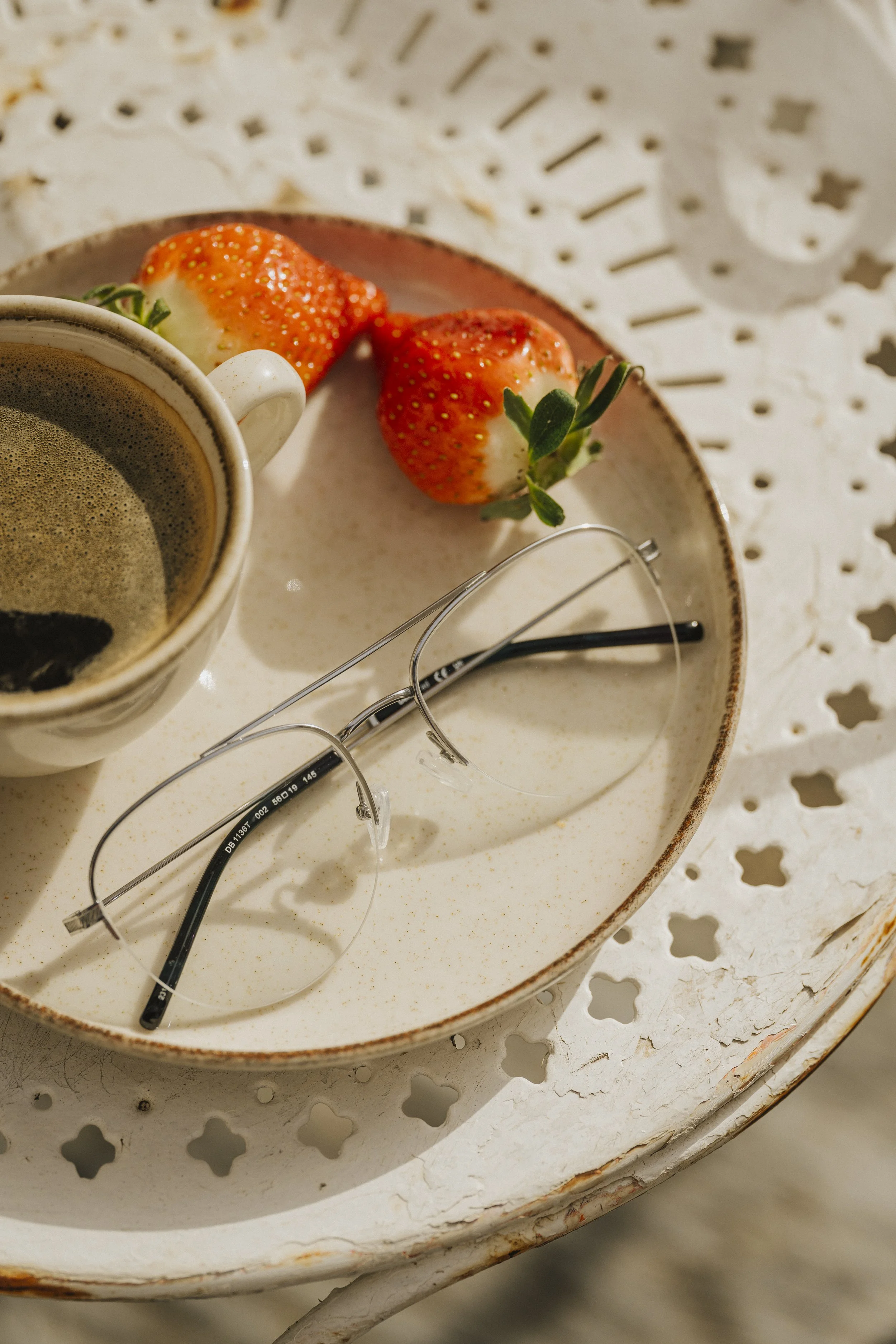 Tábua de café com duas morangos, um par de óculos de leitura e uma xícara de café, tudo sobre uma mesa de ferro com detalhes vazados.