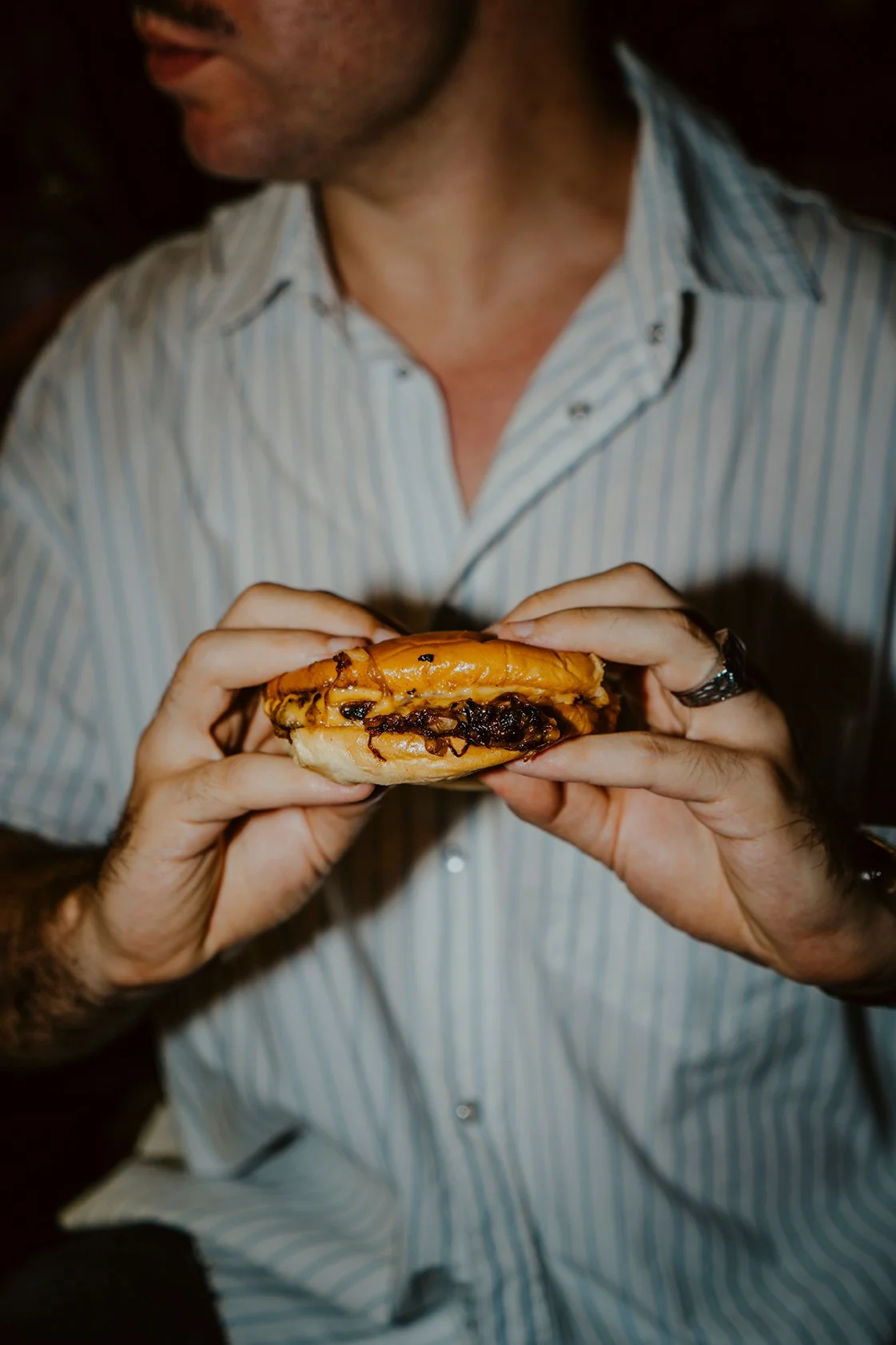 Pessoa segurando um sanduíche com queijo derretido e recheio de carne, ao usar uma camisa de mangas compridas com listras e relógio no pulso esquerdo.