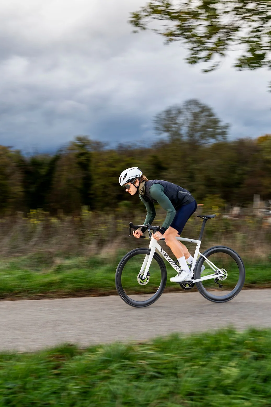 Pessoa pedalando uma bicicleta de estrada em uma pista ao ar livre, vestindo capacete e roupas esportivas, com árvores e céu nublado ao fundo.