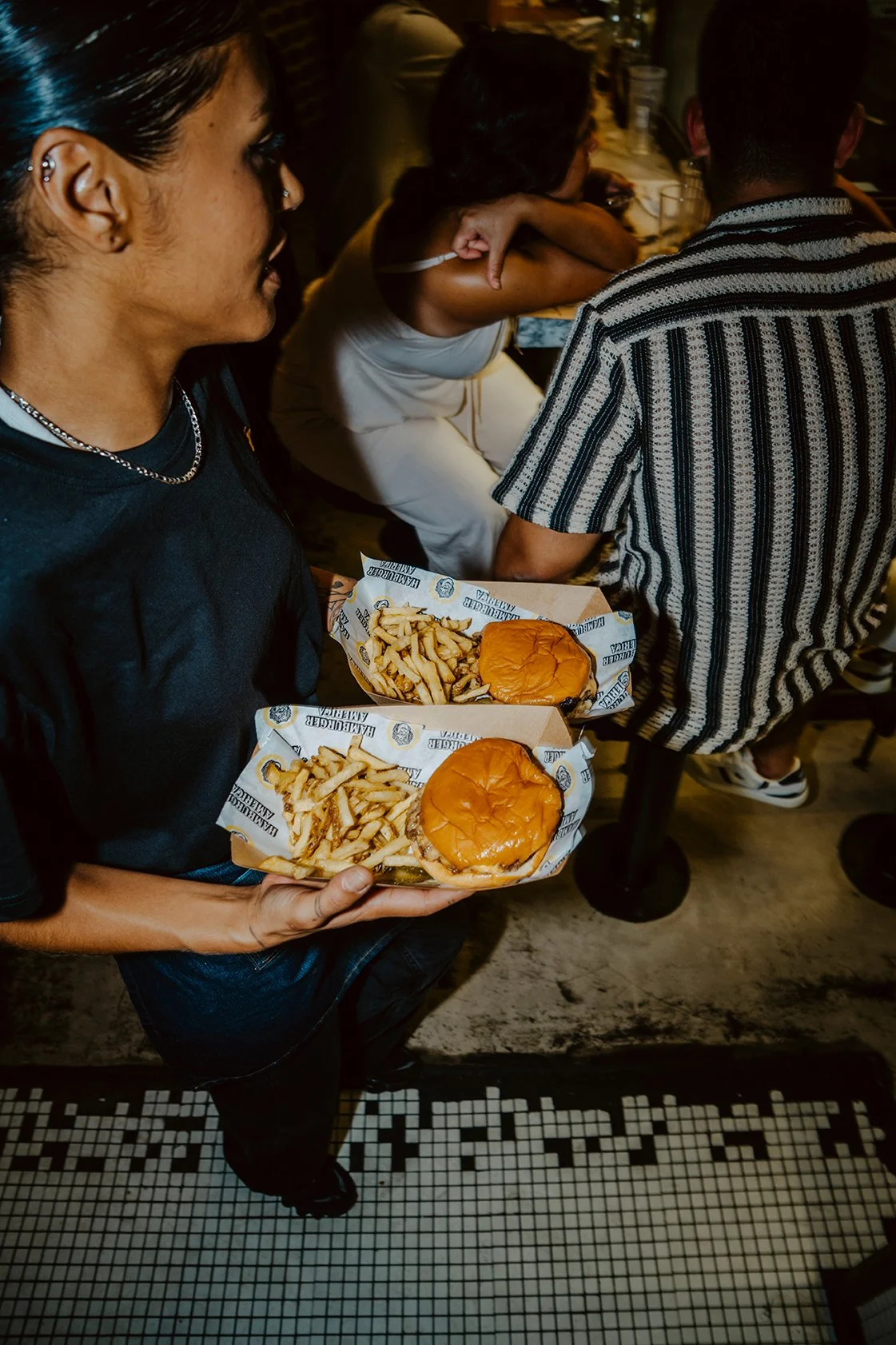 Pessoa segurando duas bandejas com hambúrgueres e batatas fritas em um restaurante movimentado.