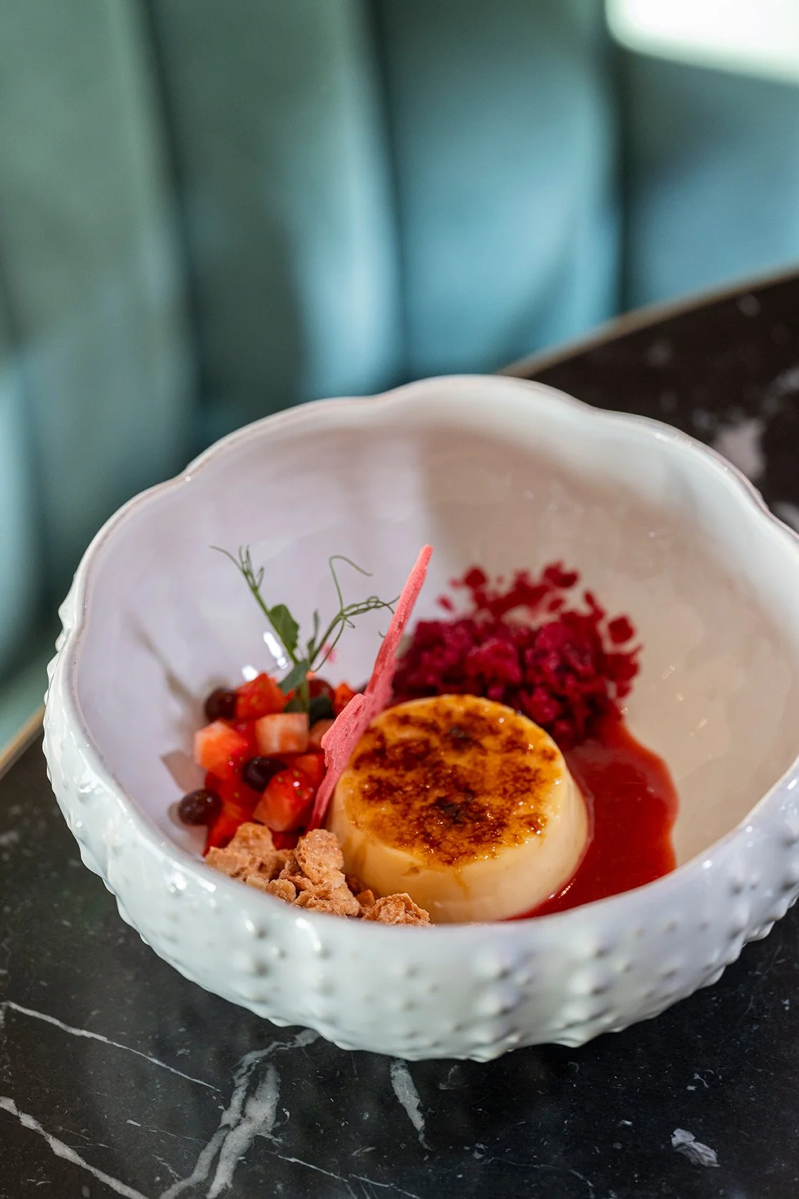 Uma taça branca com sobremesa, incluindo um creme caramelizado, frutas vermelhas, crumble e um pedaço de rosa decorativo.