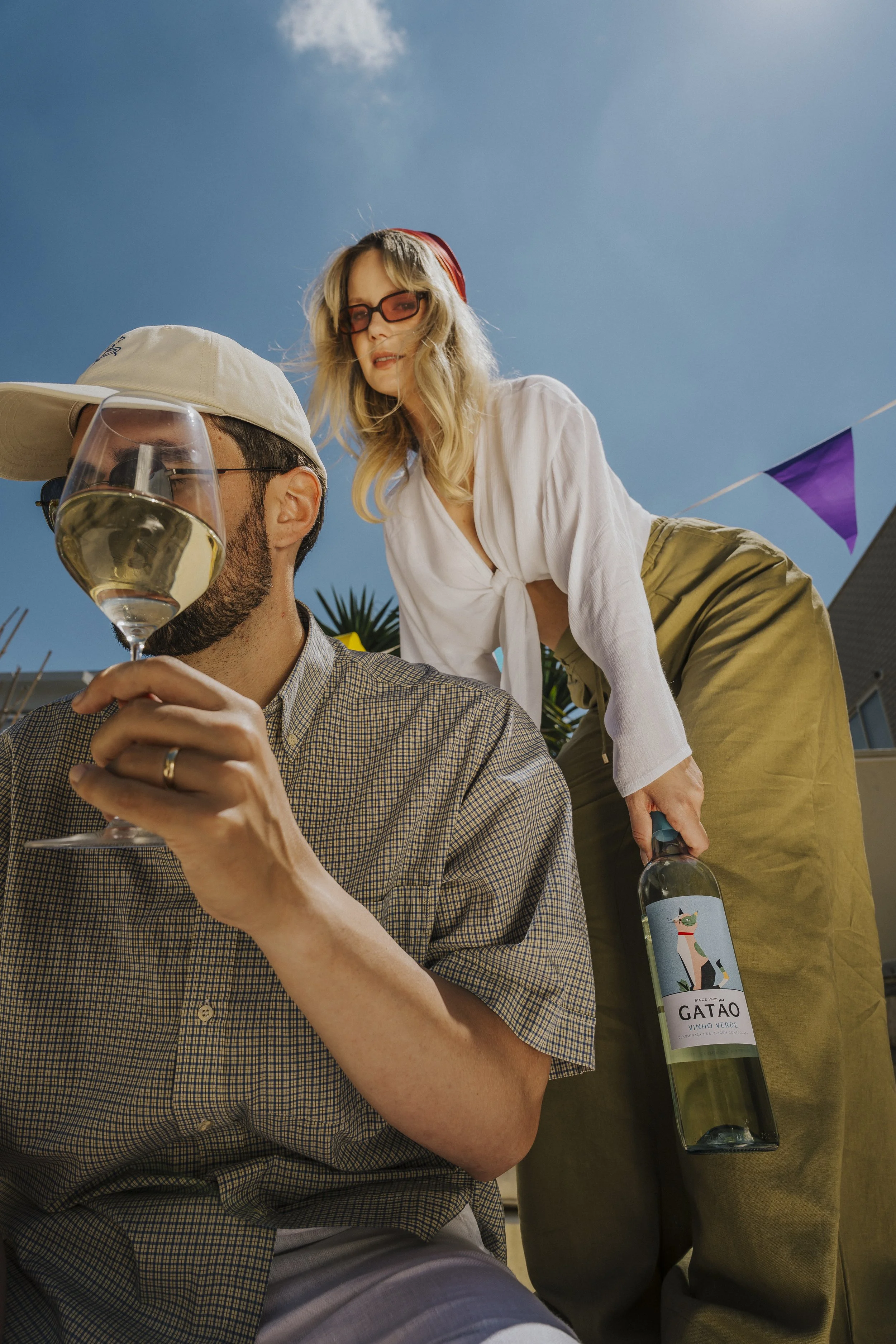 Homem com chapéu e óculos segurando uma taça de vinho branco, enquanto uma mulher ao lado dele segura uma garrafa de vinho com rótulo que diz 'Gatão Vinho Verde'. Ambos parecem estar em uma festa ao ar livre, sob céu azul, com bandeirolas decorativas.