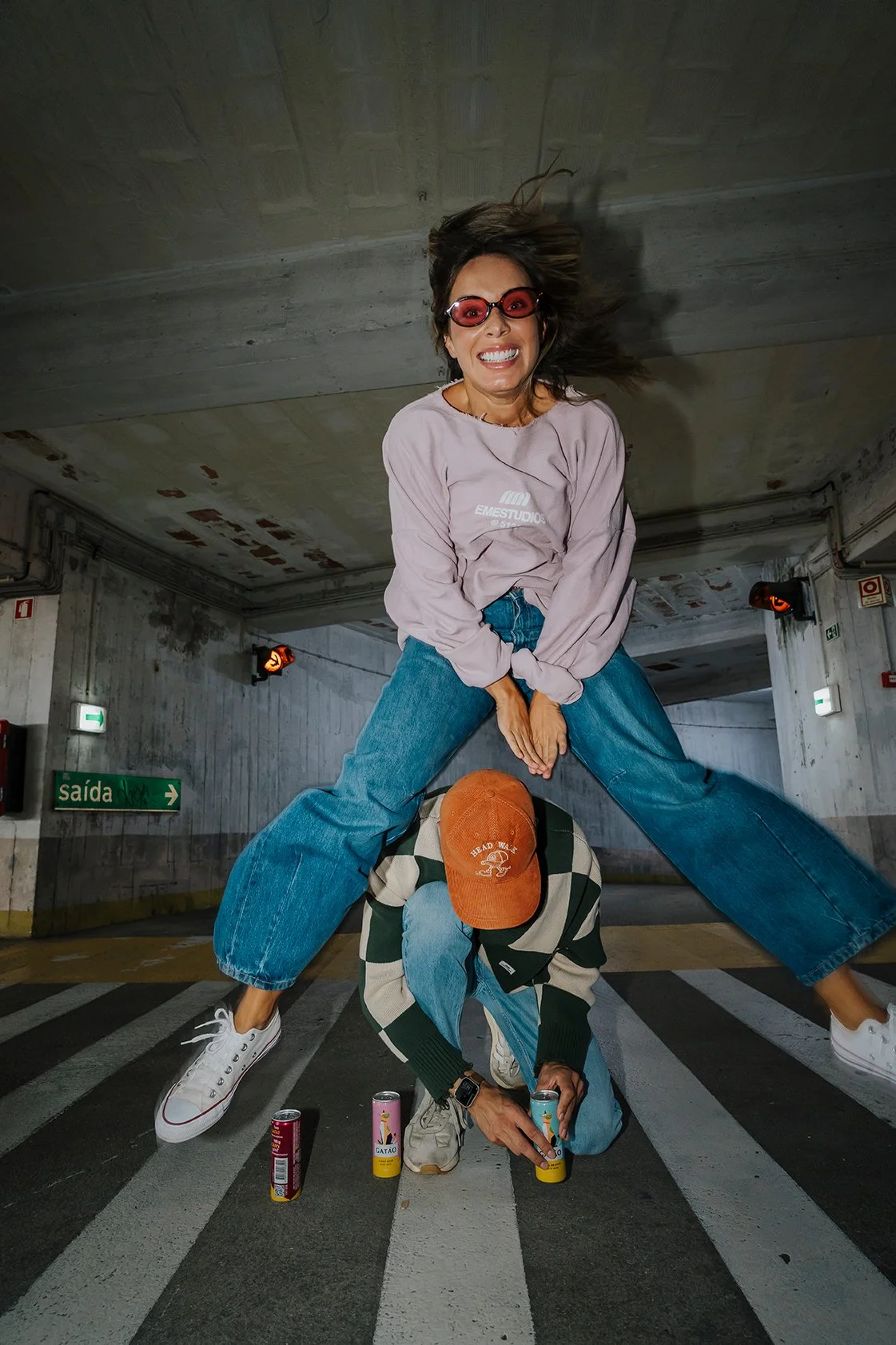 Mulher sorridente vestida com jaqueta rosa, calça jeans larga, óculos vermelhos, deitada em cima de homem de cabeça baixa usando boné laranja, casaco listrado, em uma garagem.
