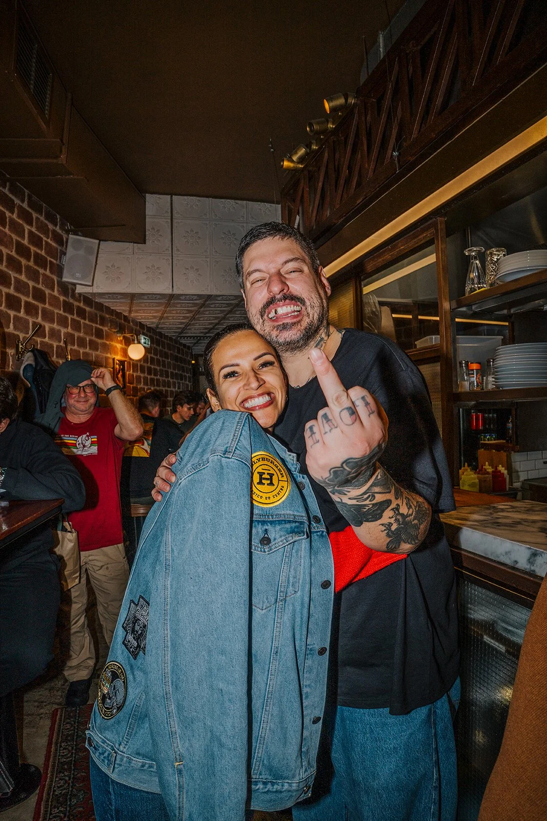 Homem e mulher sorrindo juntos em um restaurante, fazendo o gesto de middle finger, com o homem exibindo tatuagens na mão e no braço.
