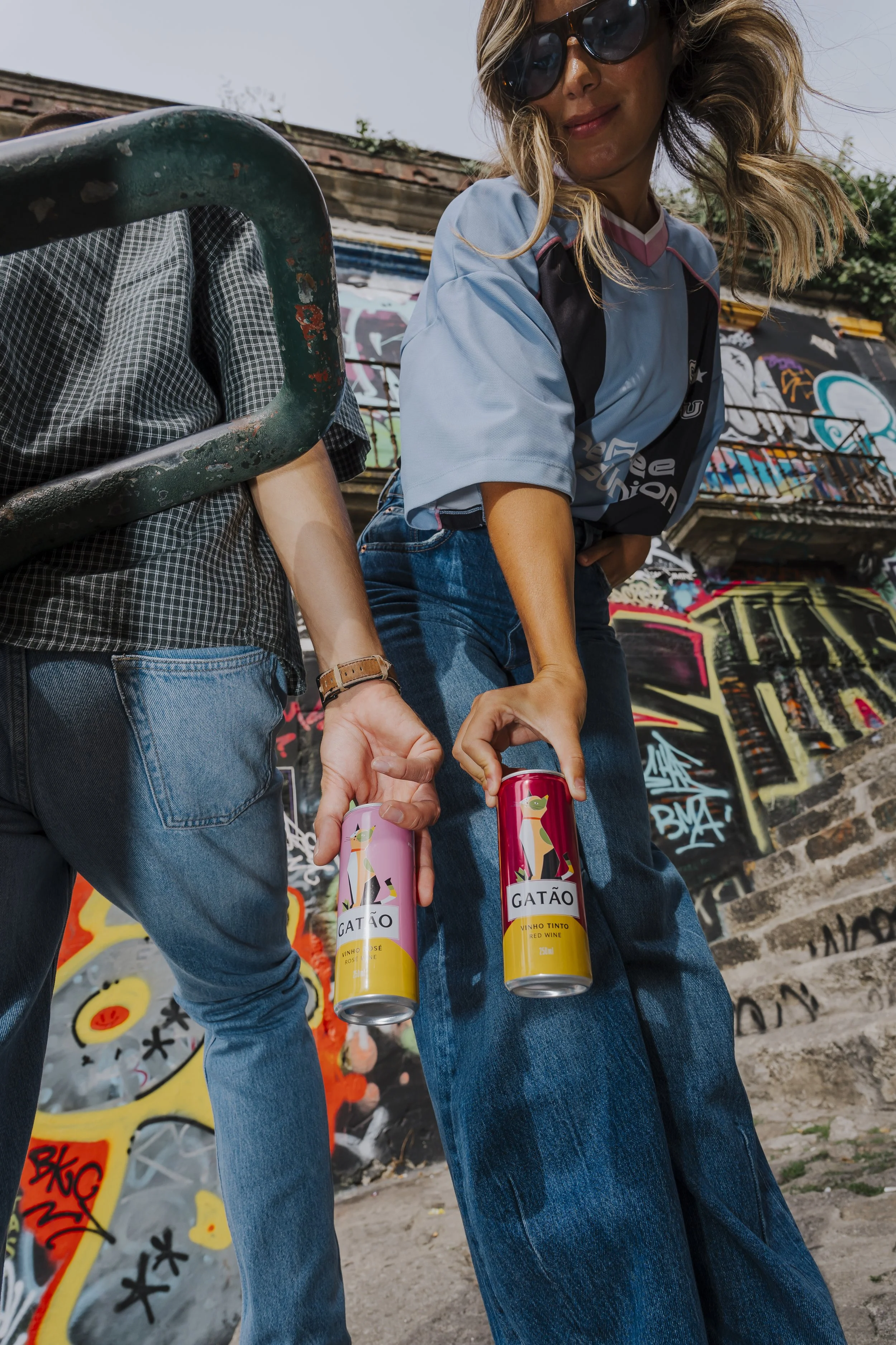 Dois jovens segurando latas de bebida com rótulo "Gatão", em frente a uma parede de graffiti, com um skate ao lado.