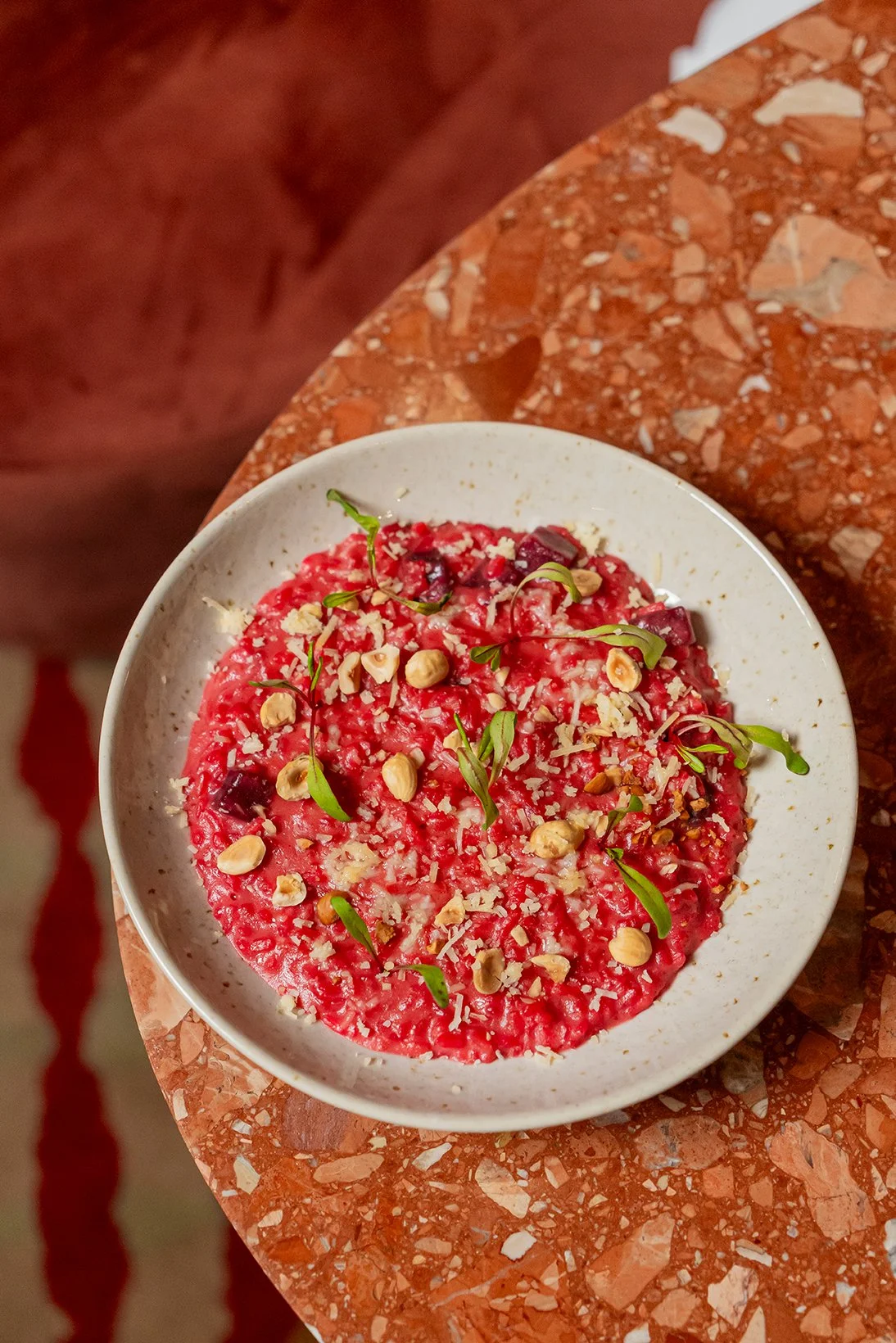 Prato de risoto de beetroot com queijo, nozes, sementes e ervas verdes, sobre mesa de mármore vermelho.