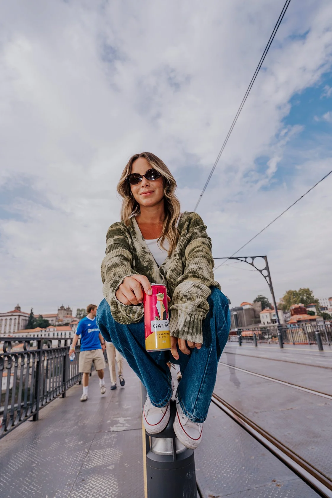 mulher sentada em uma coluna na ponte de uma cidade, segurando uma lata de vinho, usando óculos escuros, com pessoas ao fundo e céu com nuvens.