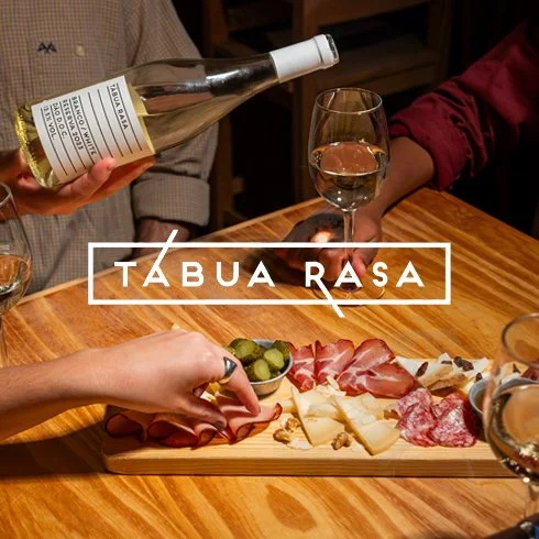 Prato com queijos, embutidos e picles, ao lado de uma pessoa servindo vinho branco em taça, em ambiente de mesa de jantar.