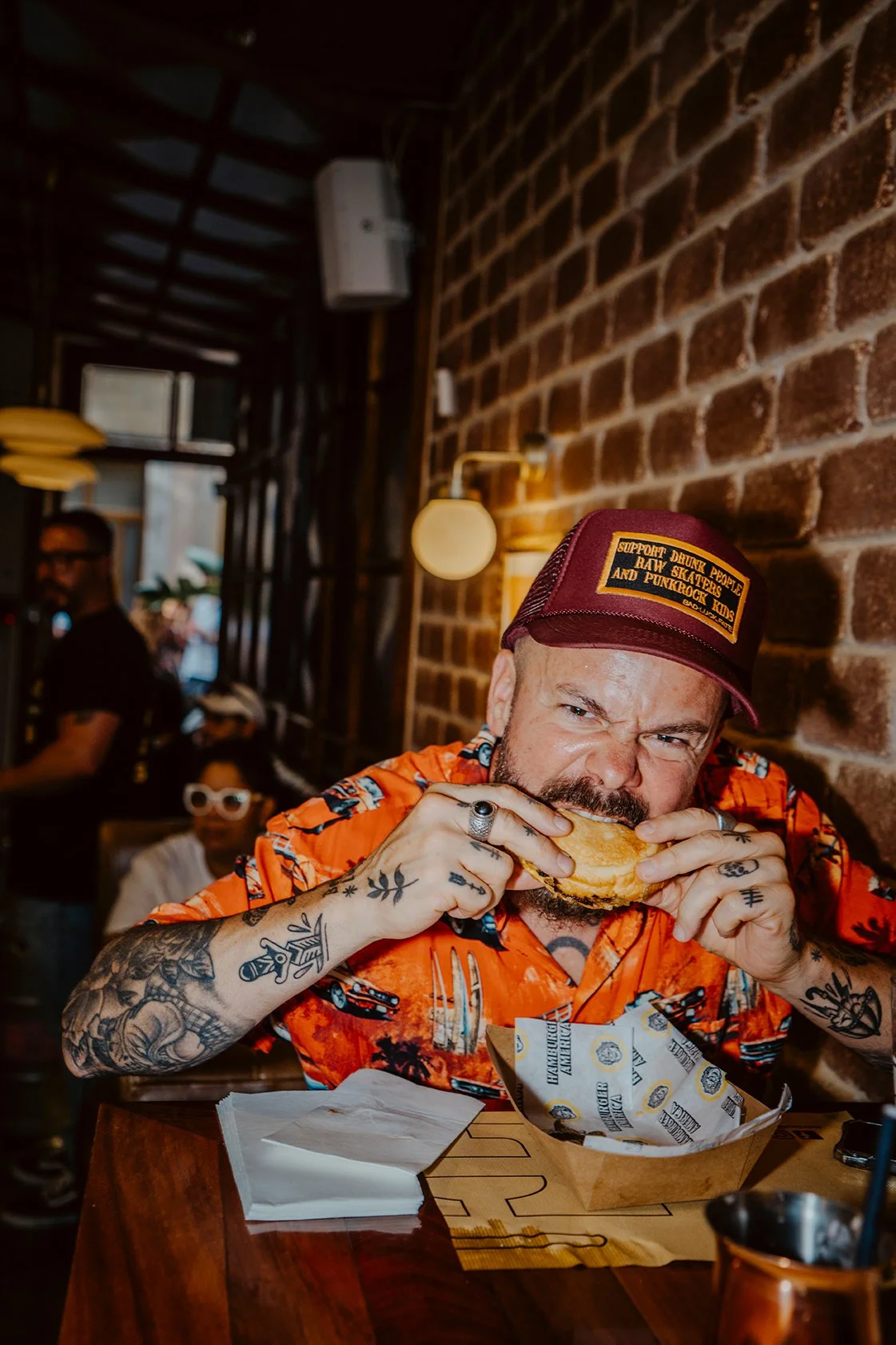Homem com tatuagens, chapéu e camisa laranja, comendo hambúrguer em restaurante com parede de tijolos expostos.