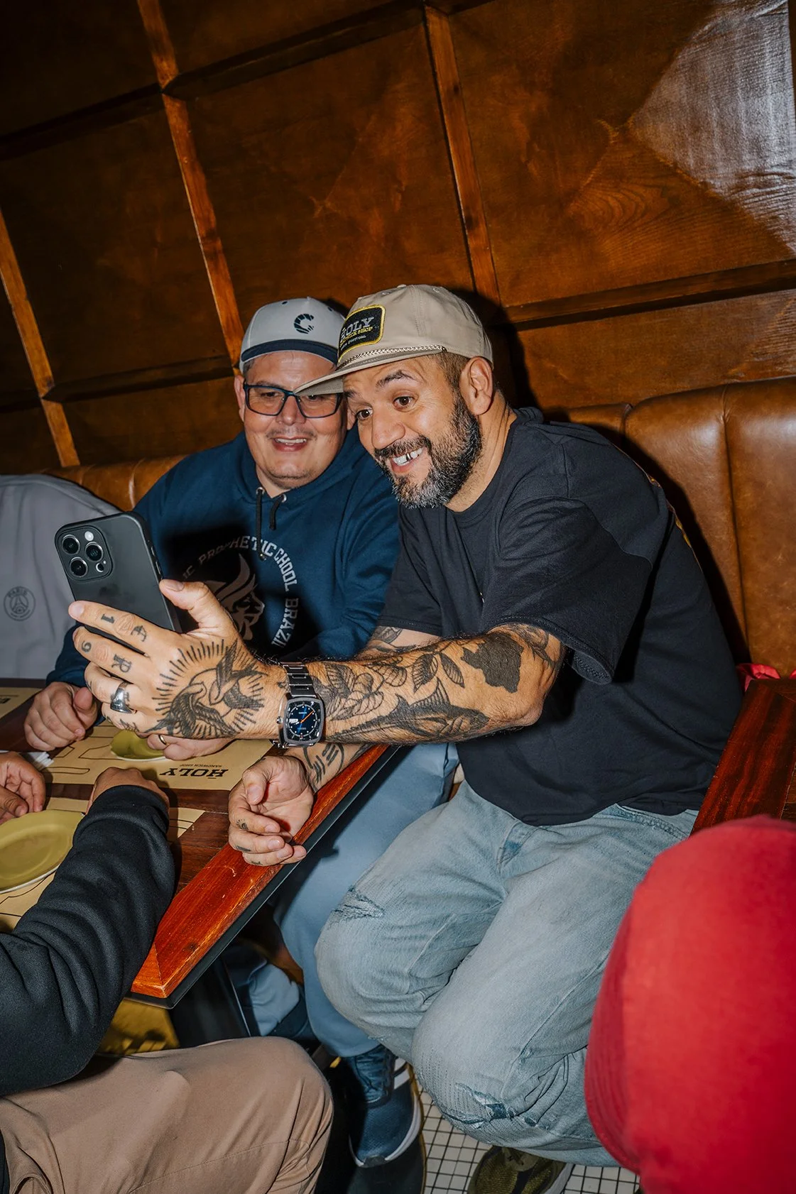 Dois homens sentados em uma mesa de restaurante, um deles usando boné e vestindo camiseta preta, ambos sorrindo enquanto olham para um celular na mão de um deles, com fundo de paredes de madeira.