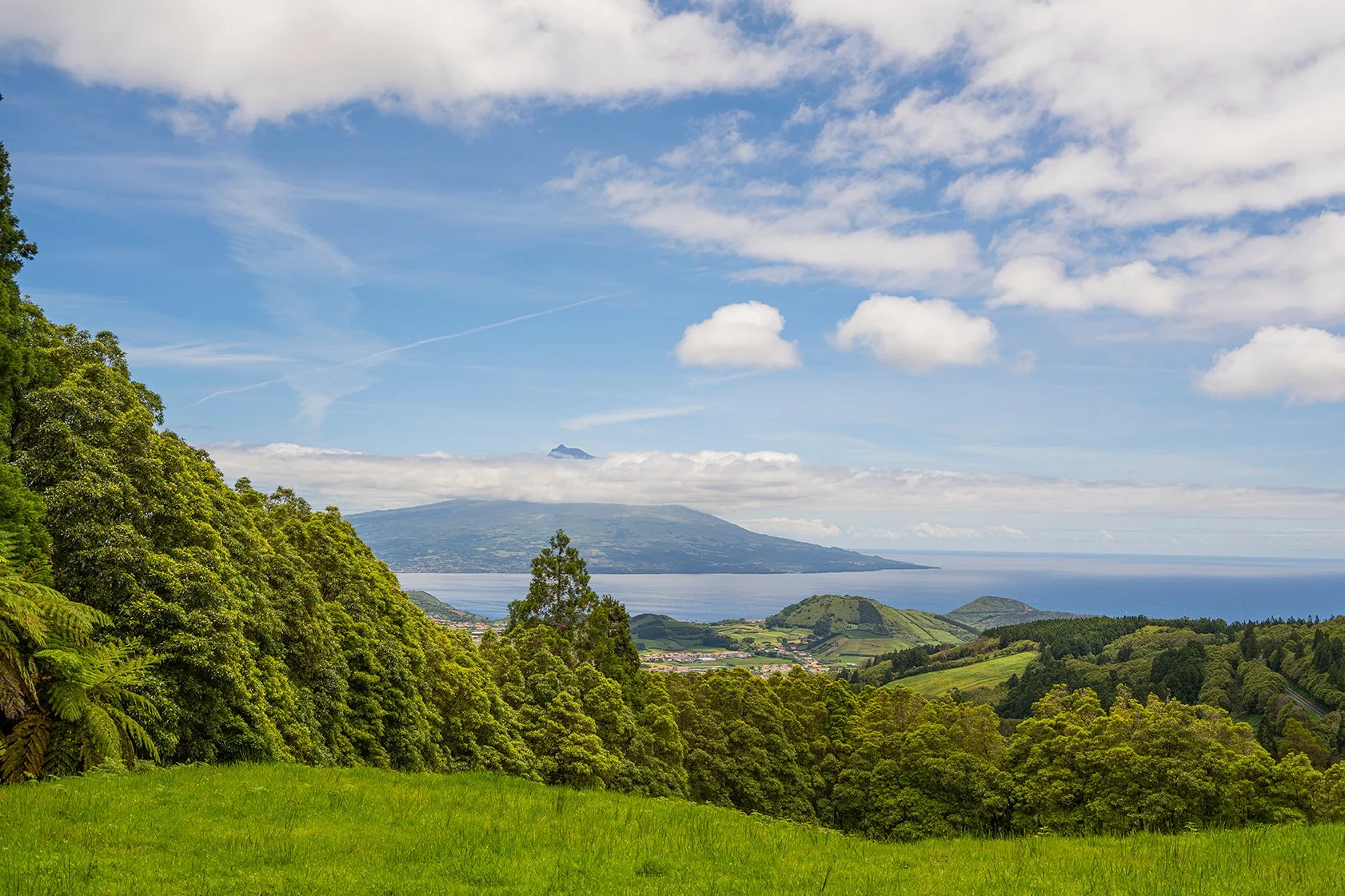 Paisagem com árvores verdes, colinas, mar e uma montanha ao fundo sob céu com nuvens.