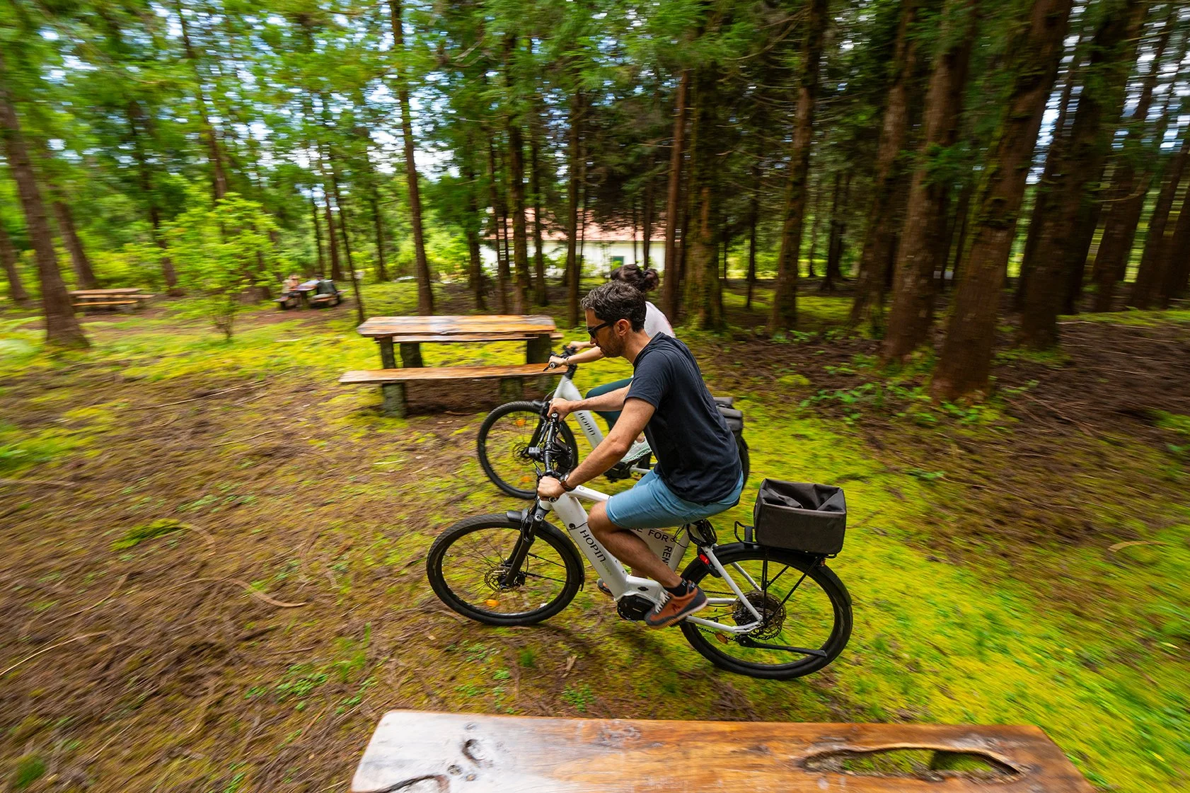 Homem e mulher pedalando em um parque de árvores verdes, com bancos de madeira ao lado.