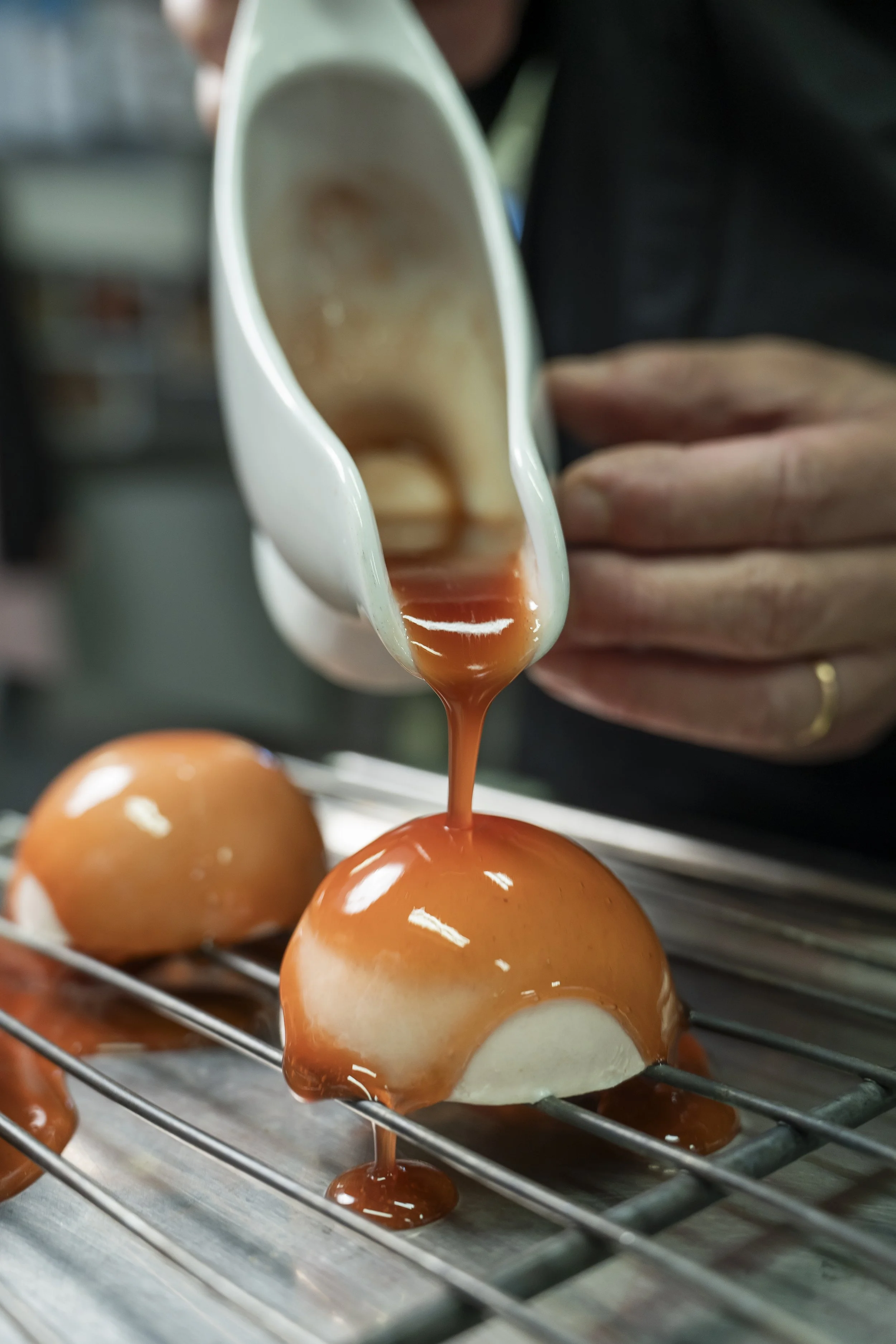 Uma pessoa despeja calda de caramelo sobre ovos de chocolate na forma de meia esfera, que estão em uma grade de refrigeração.