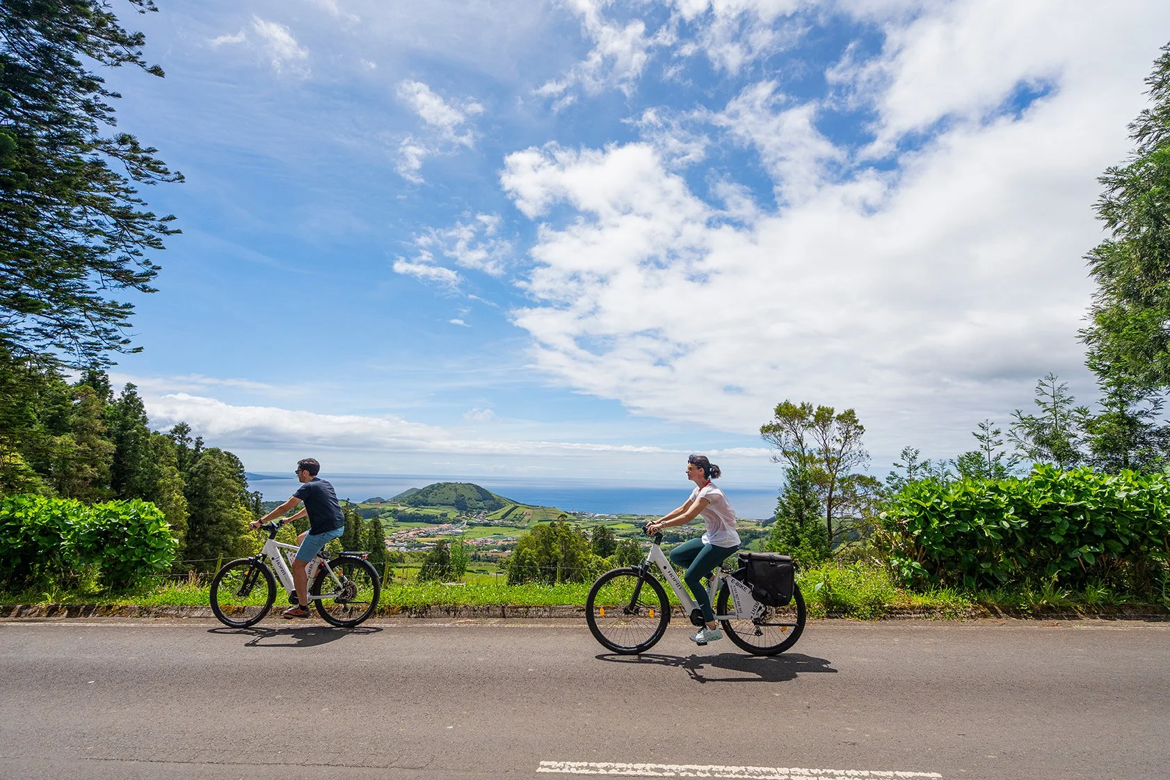 Homens e mulher pedalando de bicicleta na estrada com vista para o mar e a paisagem verde ao redor, sob céu parcialmente nublado.