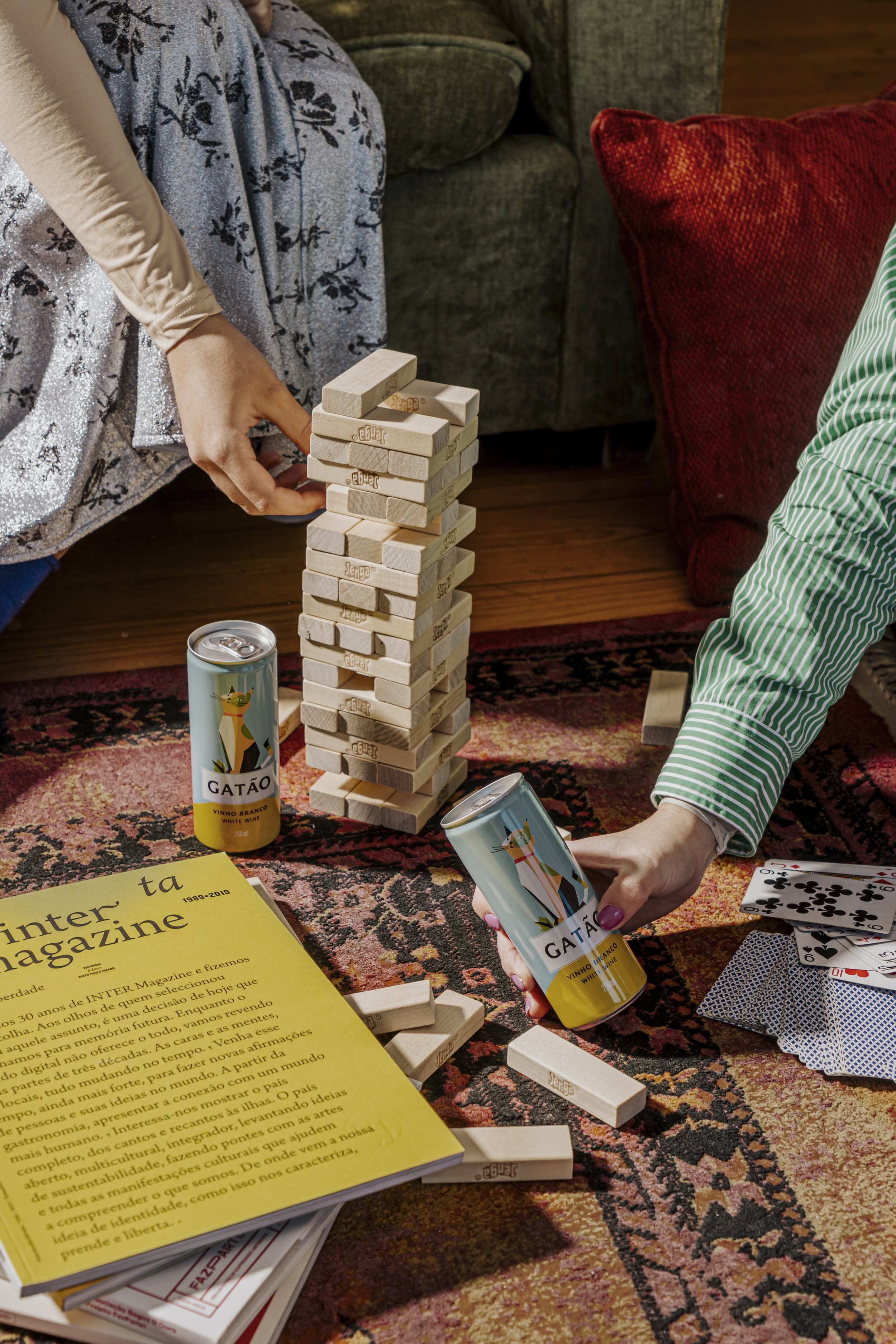Duas pessoas jogando Jenga em uma sala de estar, com uma garrafa de vinho branco GATÃO e quadrados de cartas de baralho na mesa.