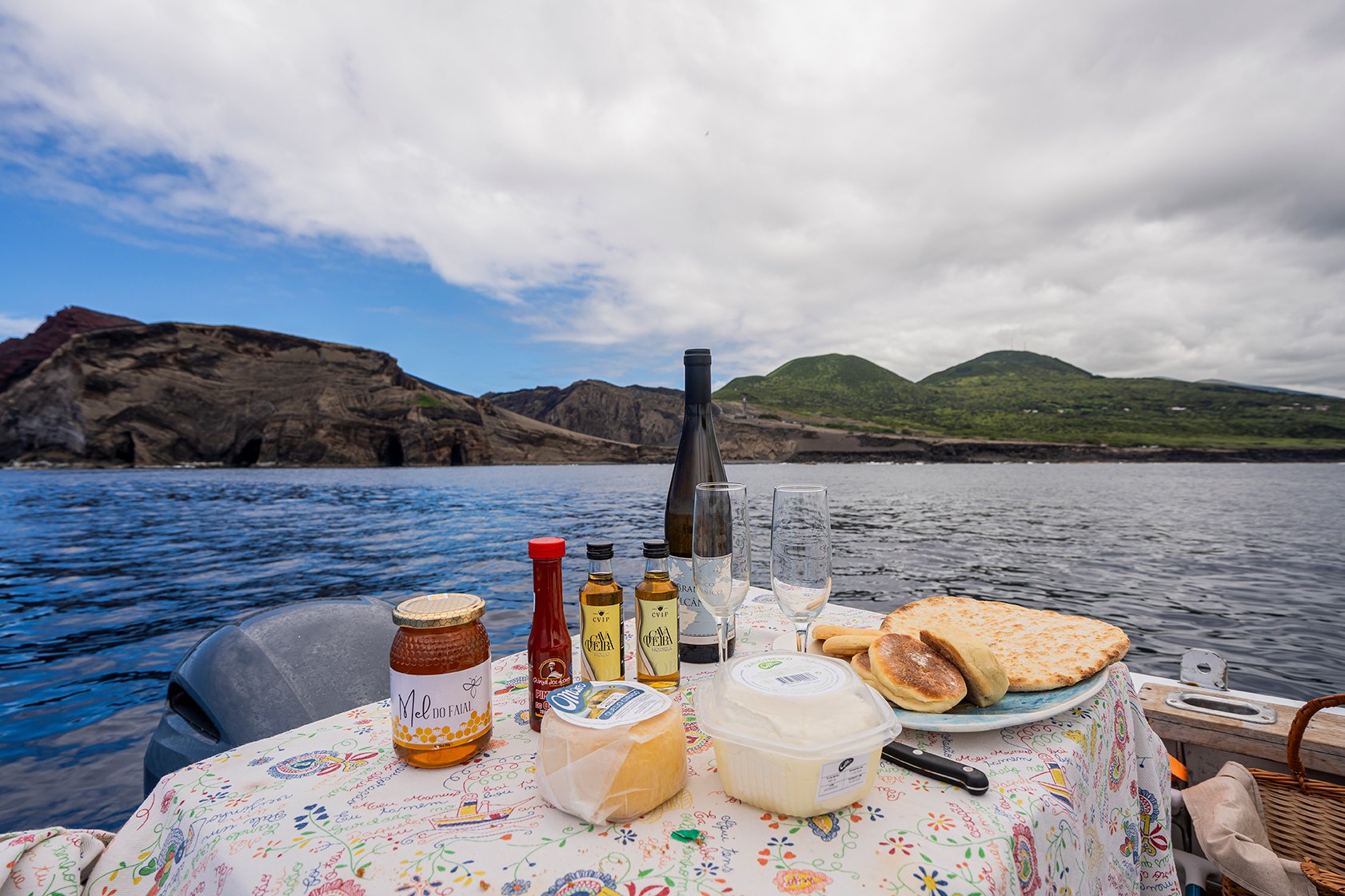 Café da manhã em um barco, com queijo, pão, geleia, bebidas, e taças, com vista para o mar e montanhas ao fundo.