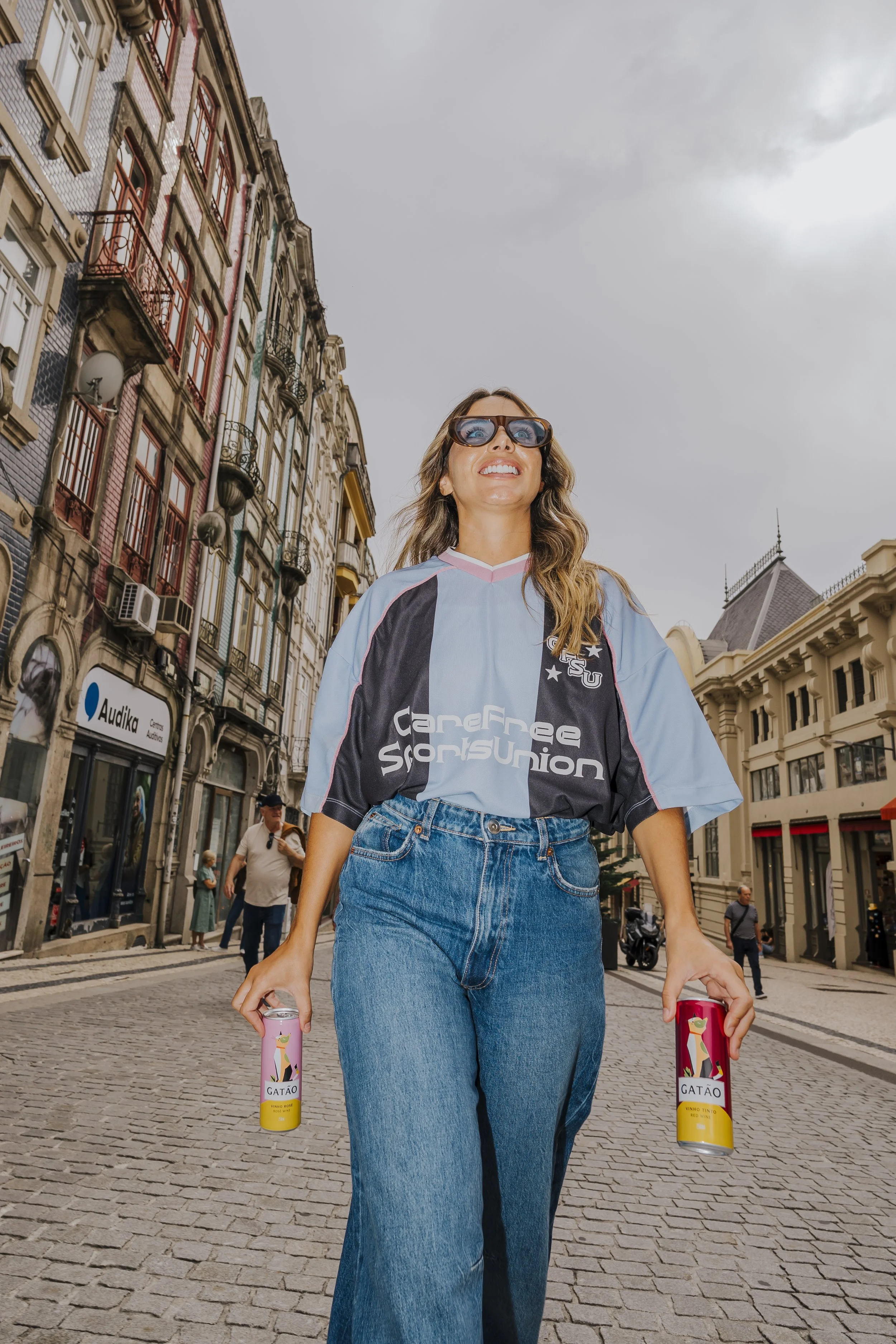 Mulher sorridente caminhando na rua de paralelepípedos, usando óculos escuros, camiseta de time esportivo azul, jeans e segurando latas de refrigerante com o rótulo 'GATÃO'.