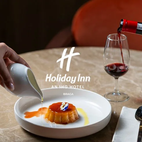 Sobremesa com pudim decorado com calda de caramelo e uma flor de confeitaria, sendo regada com leite por uma mão, com taça de vinho tinto ao lado e o logotipo do Holiday Inn Braga ao fundo.