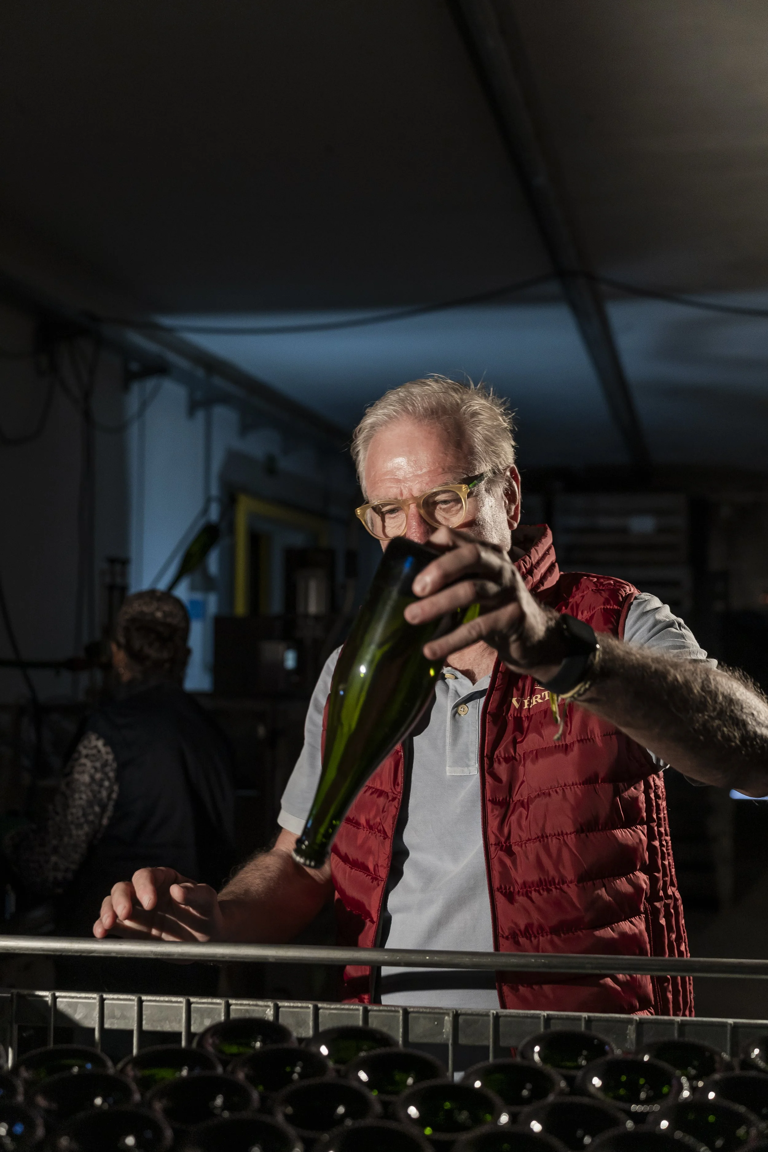 Homem com óculos de grau e cabelo grisalho, usando jaqueta vermelha, manuseando uma garrafa de vidro na frente de uma prateleira com várias garrafas de vidro, em um ambiente com iluminação baixa.