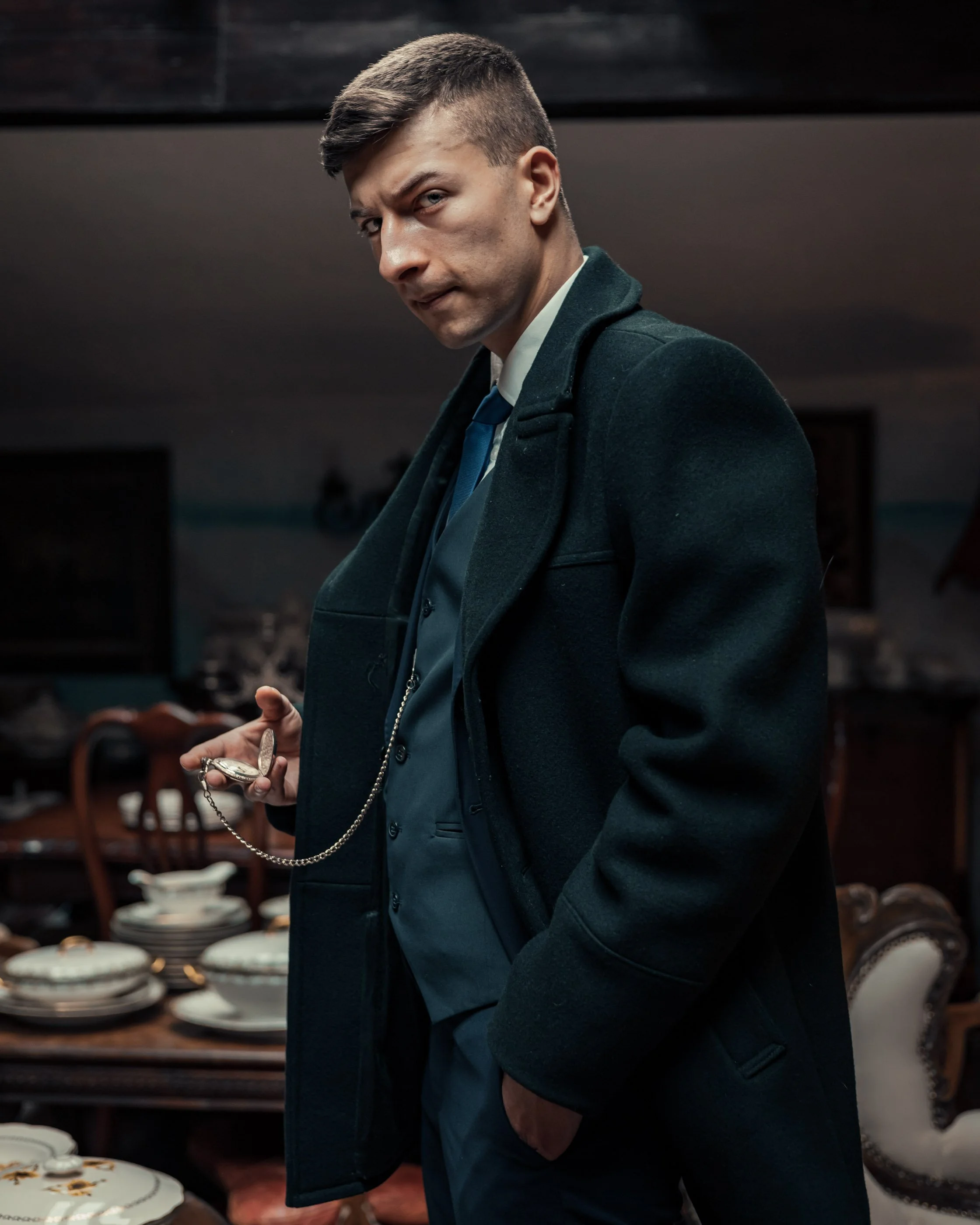 A man in a dark coat and suit holding a pocket watch with a chain in a dimly lit room with antique furniture and dishes.
