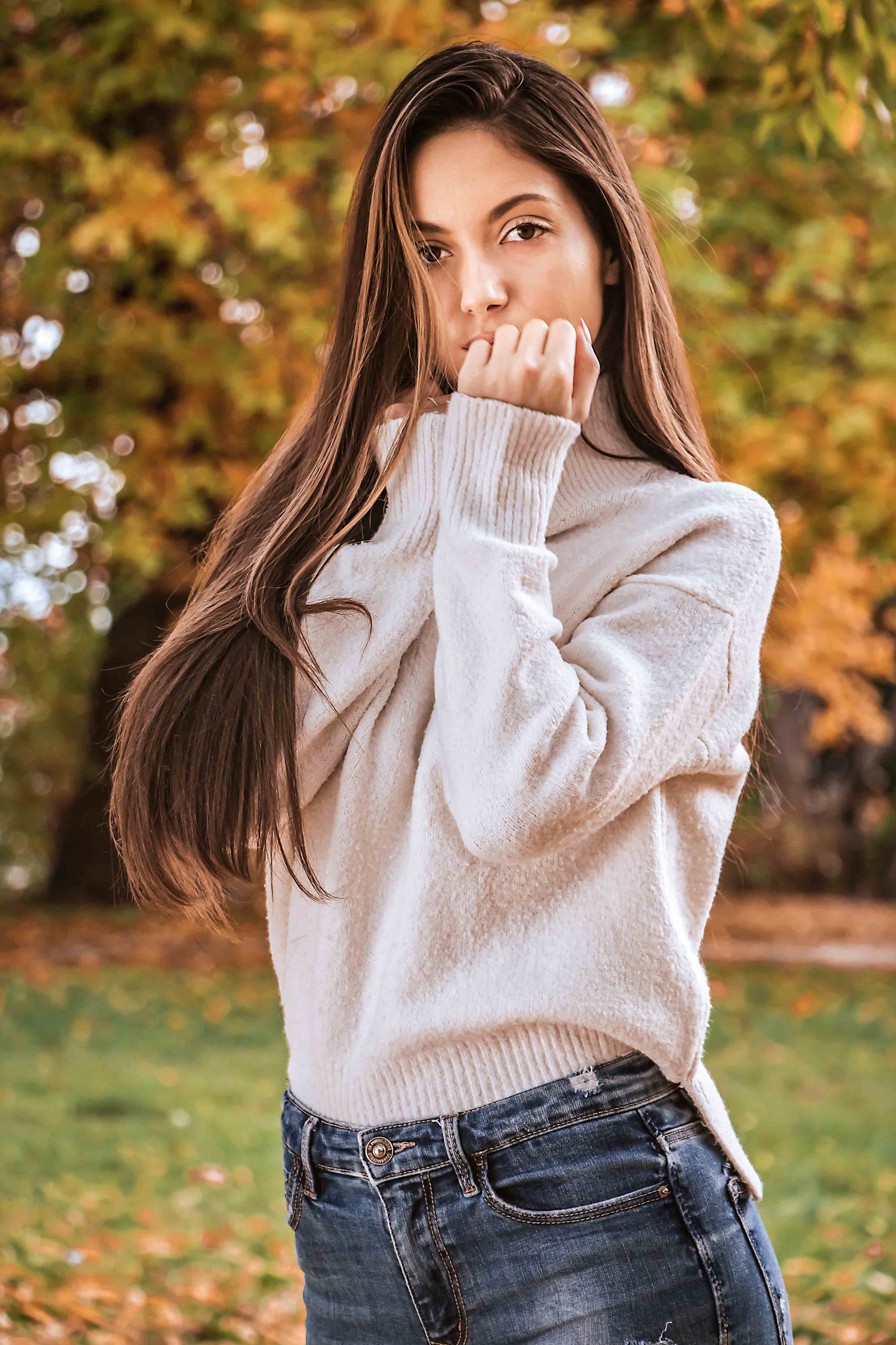 A young woman with long brown hair wearing a white sweater and dark jeans, standing outdoors in a park with autumn leaves.