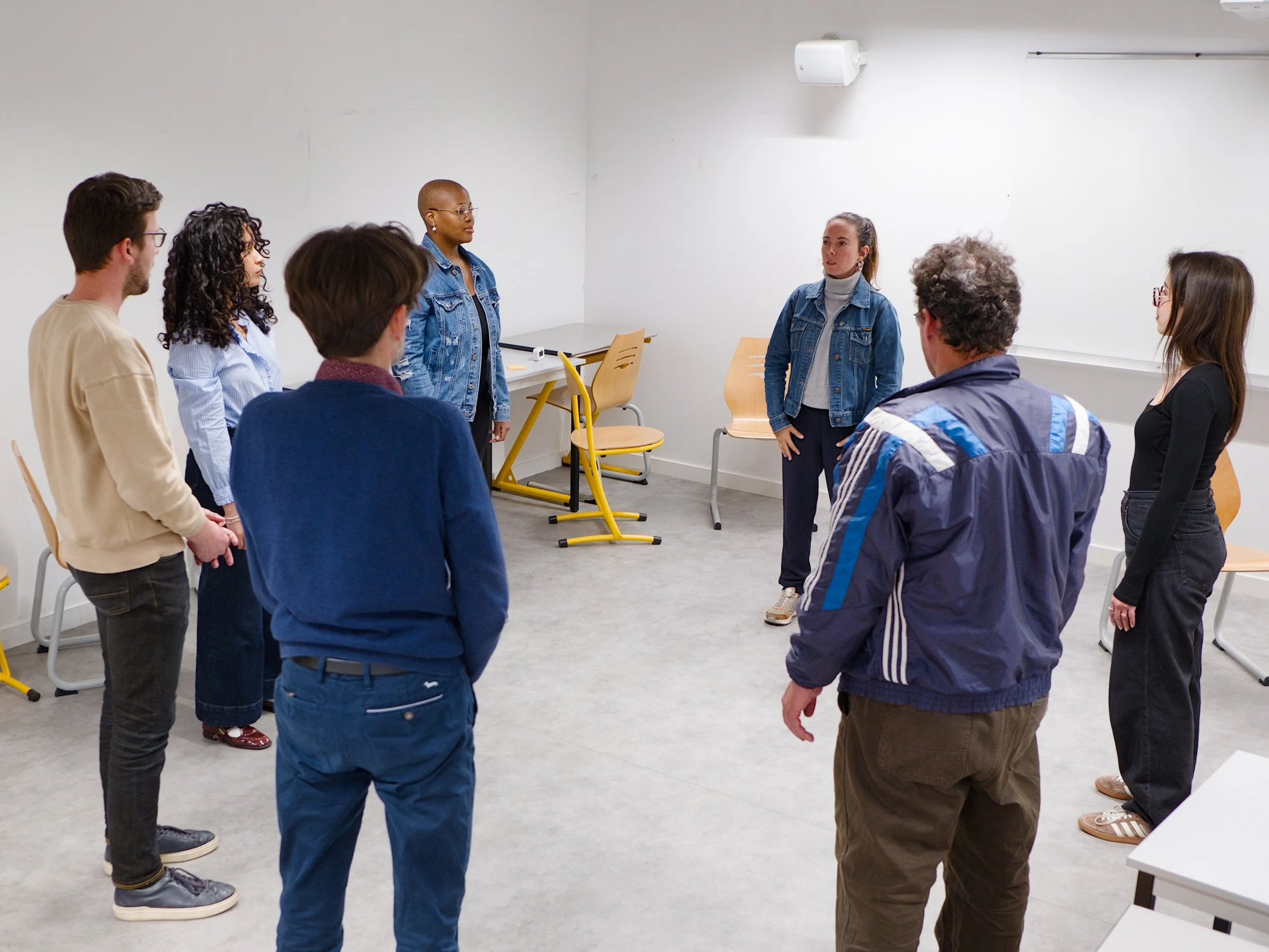 Groupe de personnes debout en cercle dans une salle blanche, deux femmes semblent discuter ou écouter tandis que d'autres personnes regardent.