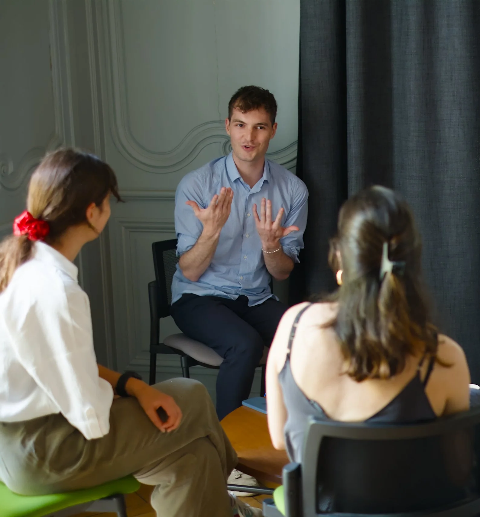 Un homme parle à un groupe de jeunes assis en cercle, dans une pièce avec murs ornés de moulures et rideaux gris, lors d'une discussion ou d'une séance de groupe.