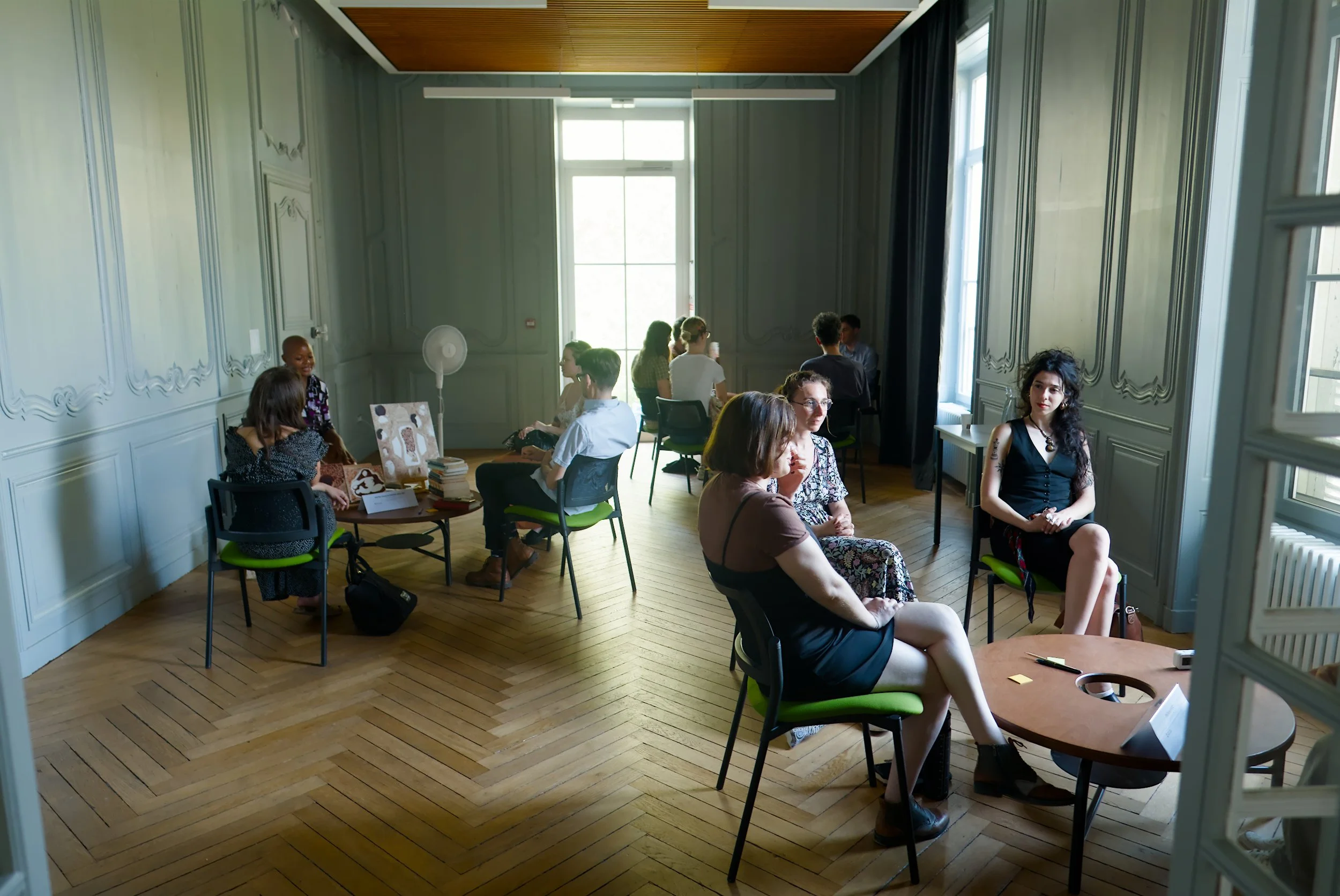 Un groupe de personnes assises dans une salle élégante avec des murs ornés, de grandes fenêtres, et des chaises vertes, semble participer à une réunion ou un atelier.