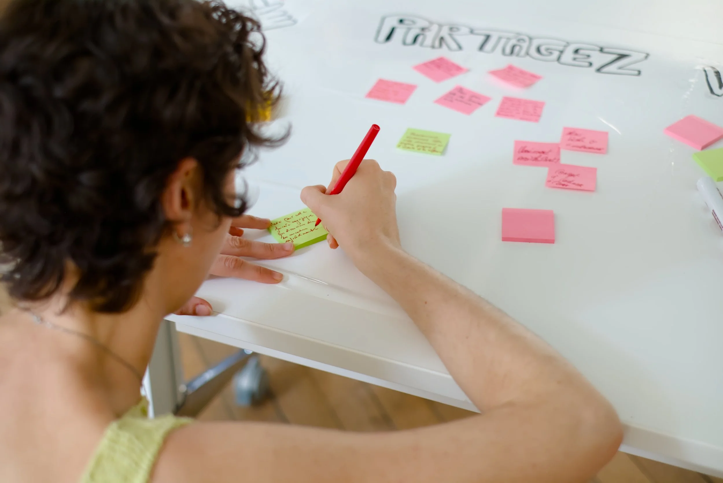 Une femme écrit sur un post-it vert avec un stylo rouge, autour des post-its roses avec des notes sur une table blanche.