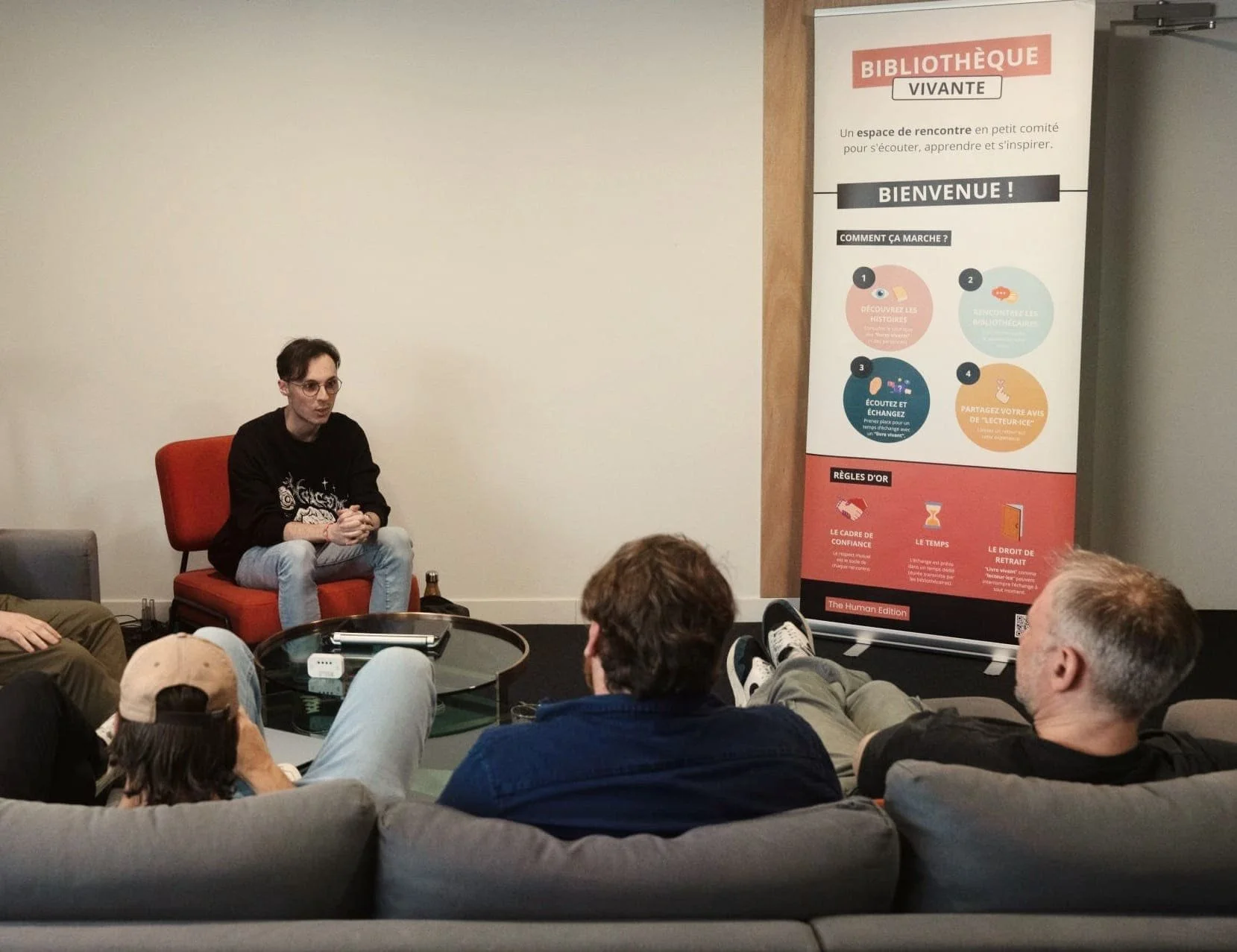 Une personne assise sur une chaise rouge donne une présentation ou intervient devant un groupe de personnes en salle, avec une bannière d'information sur le côté. La bannière décrit un espace de rencontre en bibliothèque, expliquant comment cela fonctionne et mentionne des règles d'or pour cet espace.