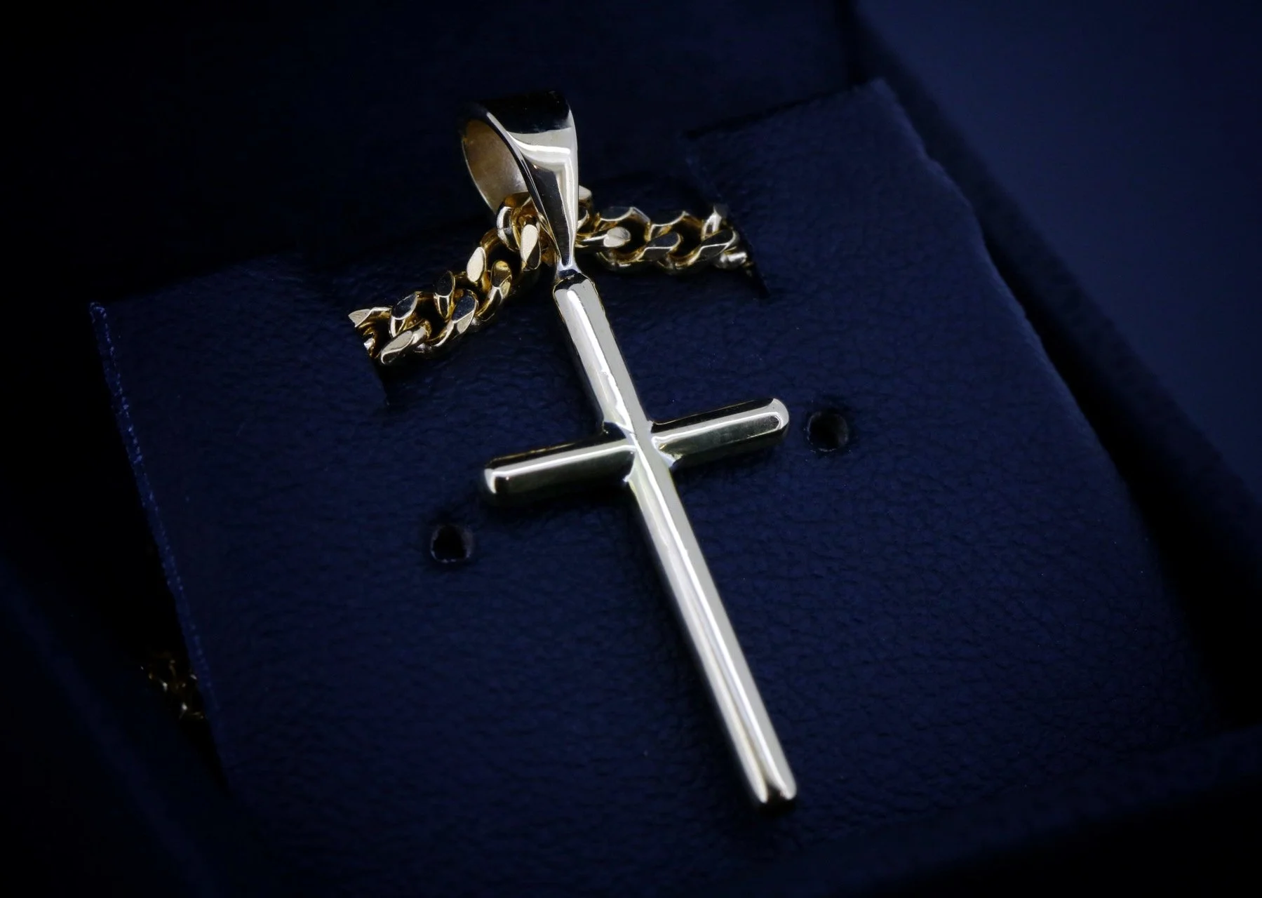 A silver cross pendant attached to a gold chain, resting on a dark textured cloth background.