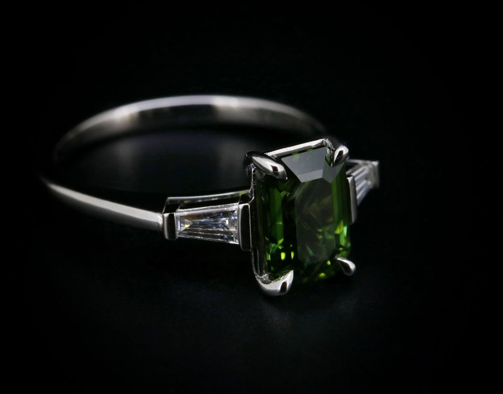 Silver ring with a large green rectangular gemstone and two smaller clear gemstones on a black background.