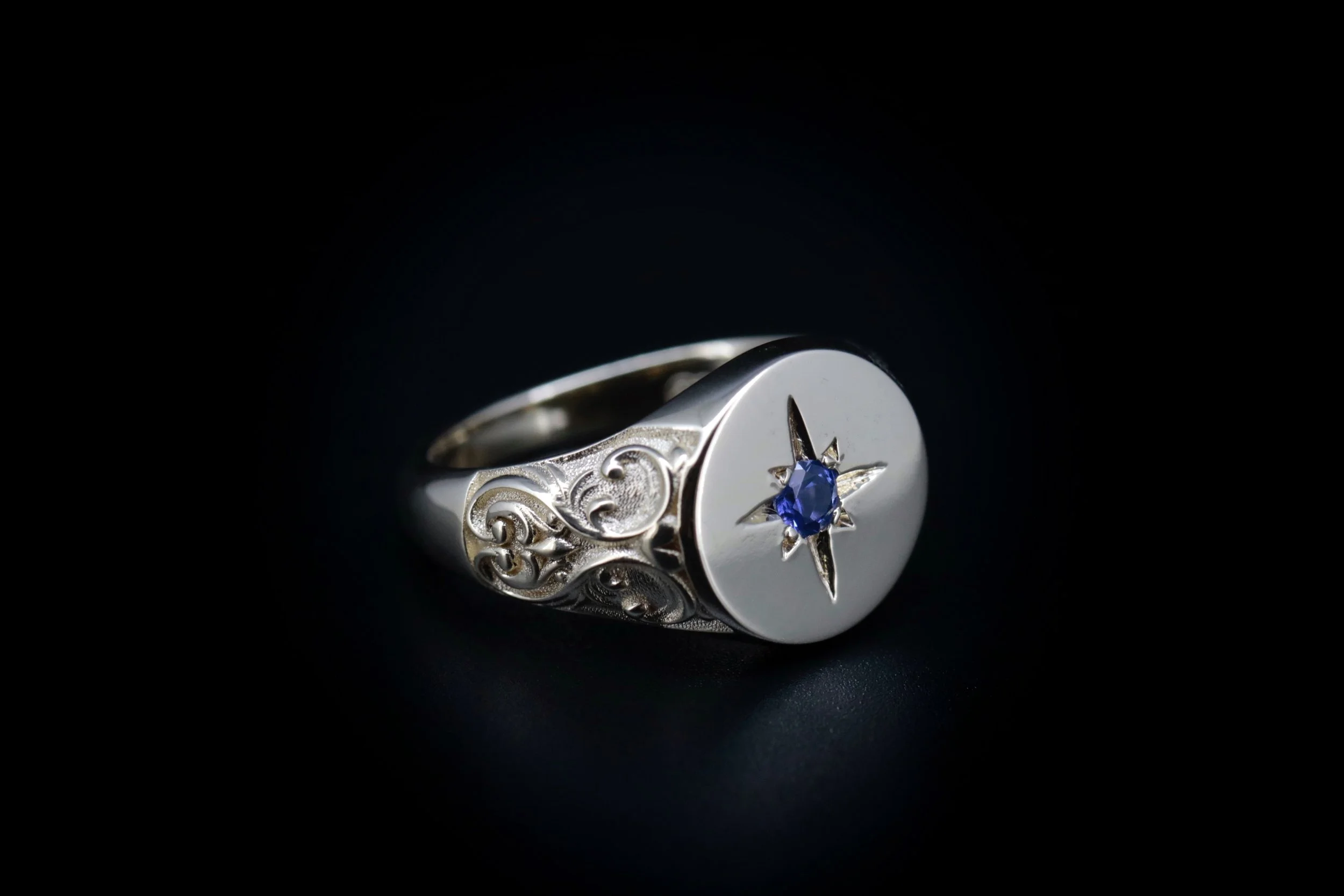 Close-up of a silver ring with a round engraved face, featuring a starburst design with a blue gemstone at the center, against a black background.