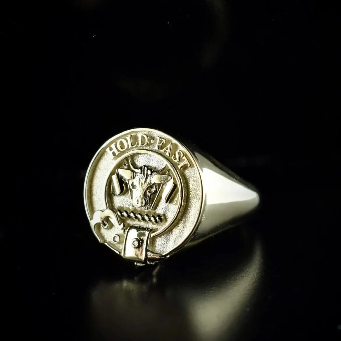 A silver ring with an engraved emblem of a bull's head and the words "HOLD. EAS" around it, set against a black background.