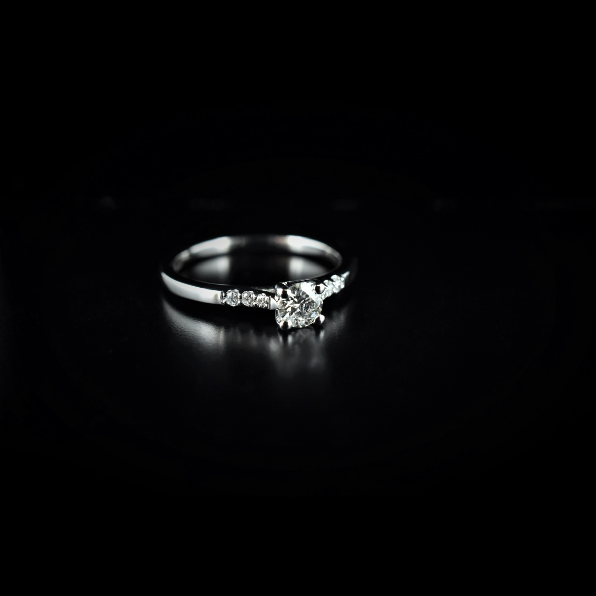 A silver engagement ring with a central round diamond and smaller diamonds on each side, set against a black background.